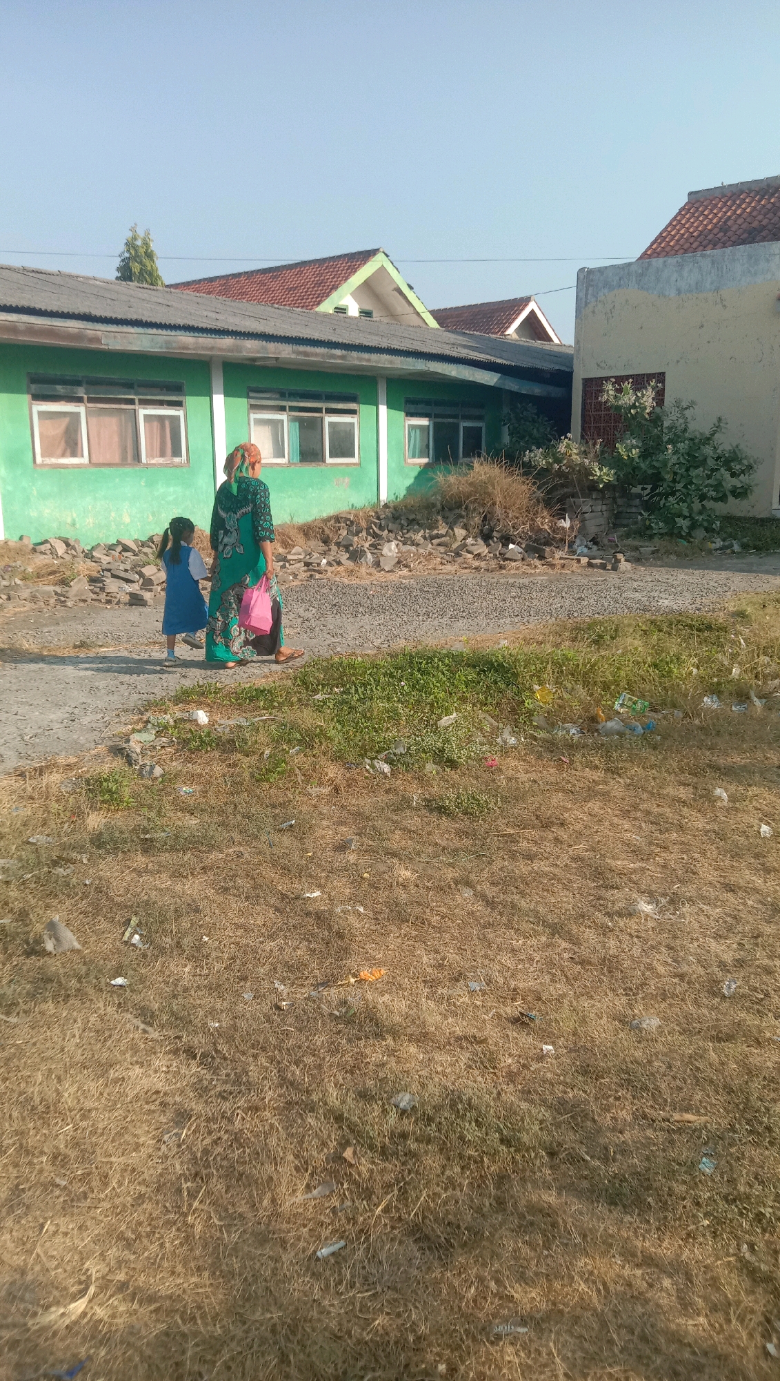 In Curah Dukuh, Indonesia, a woman in a green patterned dress and headscarf walks hand-in-hand with a young girl wearing a blue dress. They are on a dirt path in front of a long, green building with multiple windows. To the right of the building, there is a patch of dry, overgrown grass with scattered debris, including plastic bottles and other trash. Further to the right, another building with a light-colored facade and a red-tiled roof is visible. The sky is clear and blue.