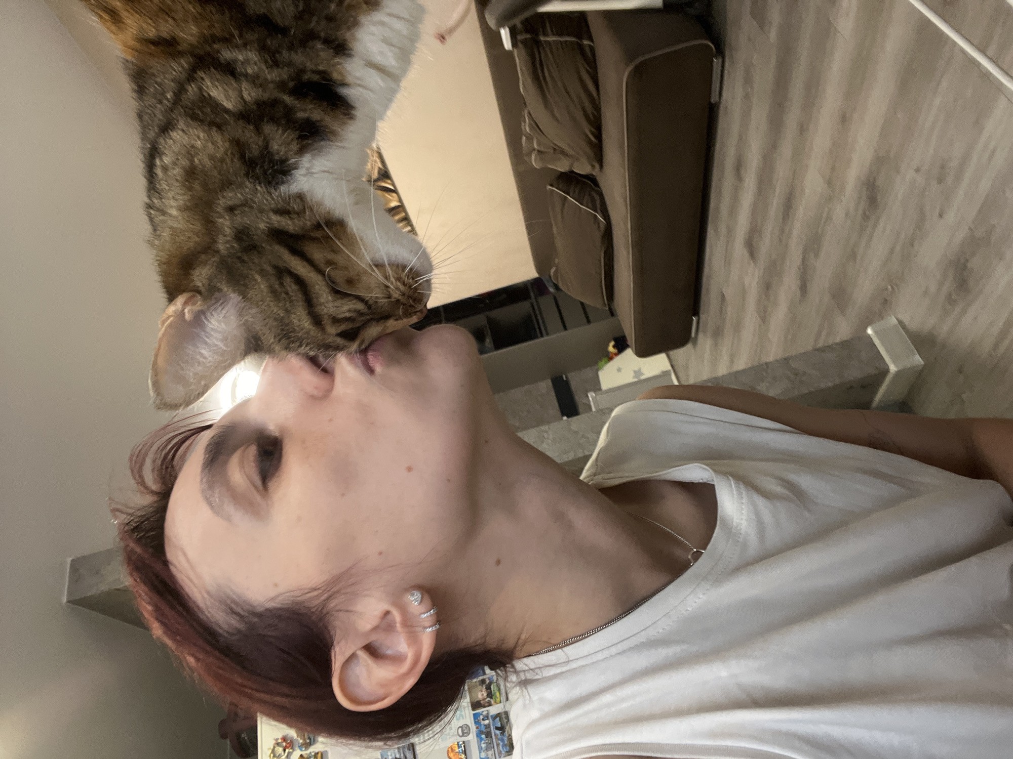 The image captures a close-up of a young woman with short, reddish-brown hair, wearing a white sleeveless shirt and a silver necklace, leaning in to kiss a tabby cat. The cat, predominantly brown and white with distinct stripes, is positioned above the woman, with its head tilted towards her. The woman has small hoop earrings and a delicate silver stud earring in her left ear. Her facial expression is one of affection. In the background, a brown sofa with two pillows is visible, along with a light-colored wall and a wooden floor. The environment appears to be an indoor residential setting. The image does not provide any specific visual cues to indicate its location within Nizhny Novgorod, Russia.