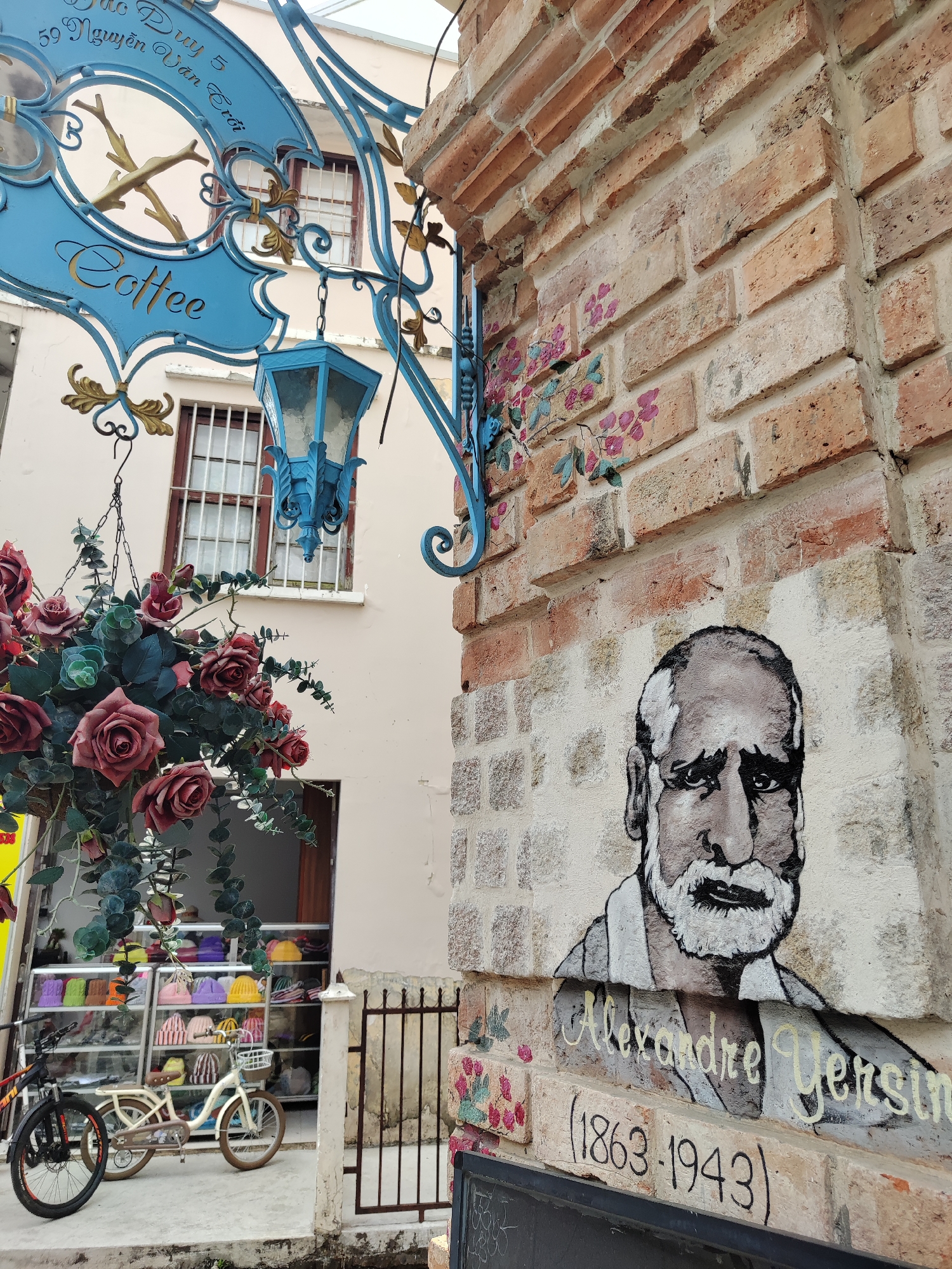 The image captures an outdoor scene in Da Lat, Vietnam, featuring a corner brick wall adorned with a painted portrait of Alexandre Yersin, along with his birth and death years, 1863-1943. Floral designs are also painted on the brick wall. A blue wrought-iron sign for "Coffee" hangs above. Hanging below the sign are vibrant faux roses and other greenery.
A storefront with a display of colorful hats and bags can be seen in the background, along with two bicycles parked in front. The building is painted a neutral color, and there's a window with bars visible in the background. A black iron fence stands adjacent to the brick wall.