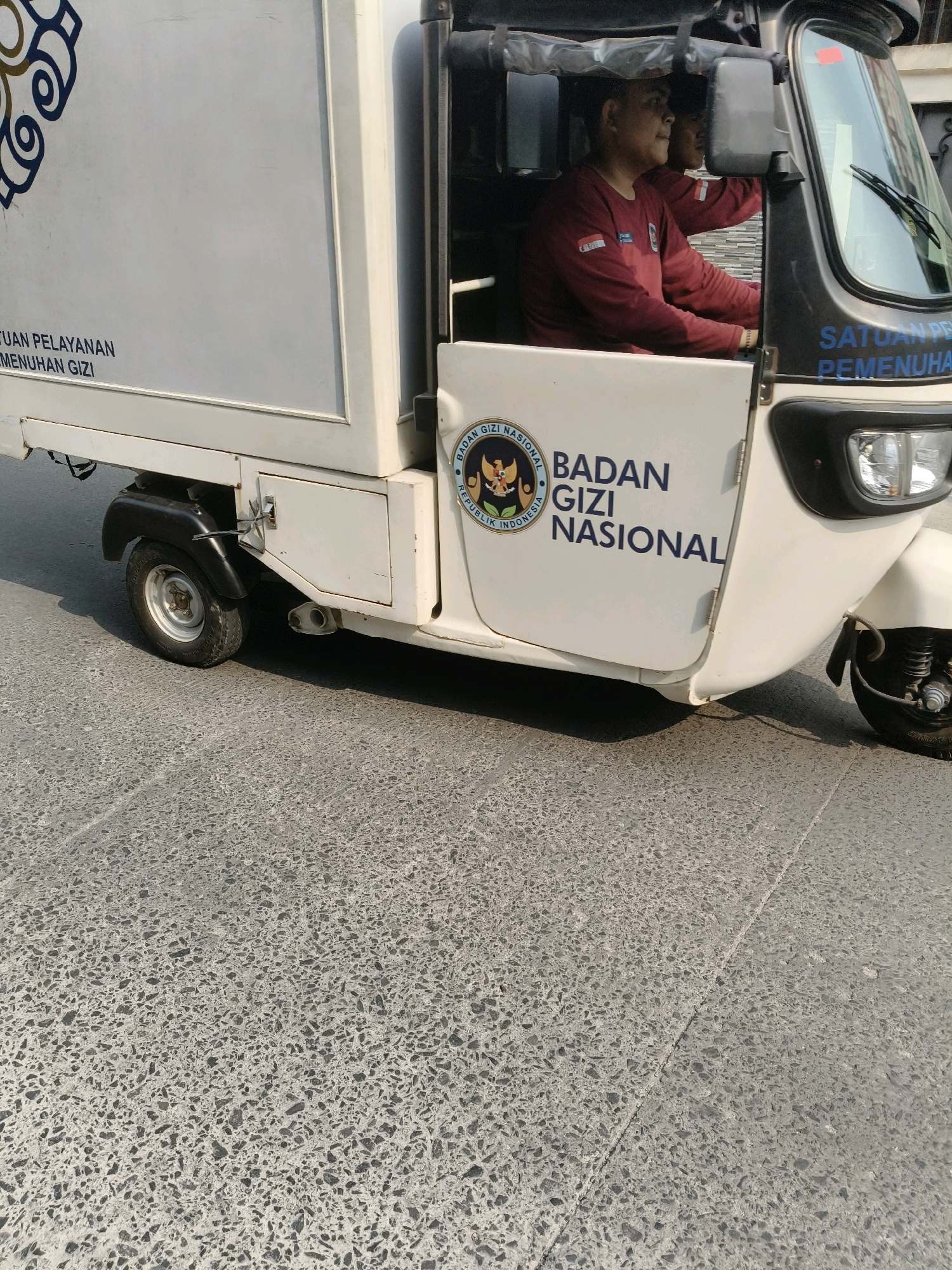 A white three-wheeled vehicle, possibly a modified tuk-tuk, is parked on a textured gray asphalt road in South Tangerang, Indonesia. The vehicle has a large white cargo box on the back and a driver's cab in the front. A man wearing a maroon long-sleeved shirt is seated in the driver's seat, looking to the right. The side of the cargo box features text in blue, including "BADAN GIZI NASIONAL" and an emblem with an eagle and text that appears to be in Indonesian. A logo in the upper left corner of the cargo box is partially visible. The scene is lit by sunlight, casting shadows on the road.
