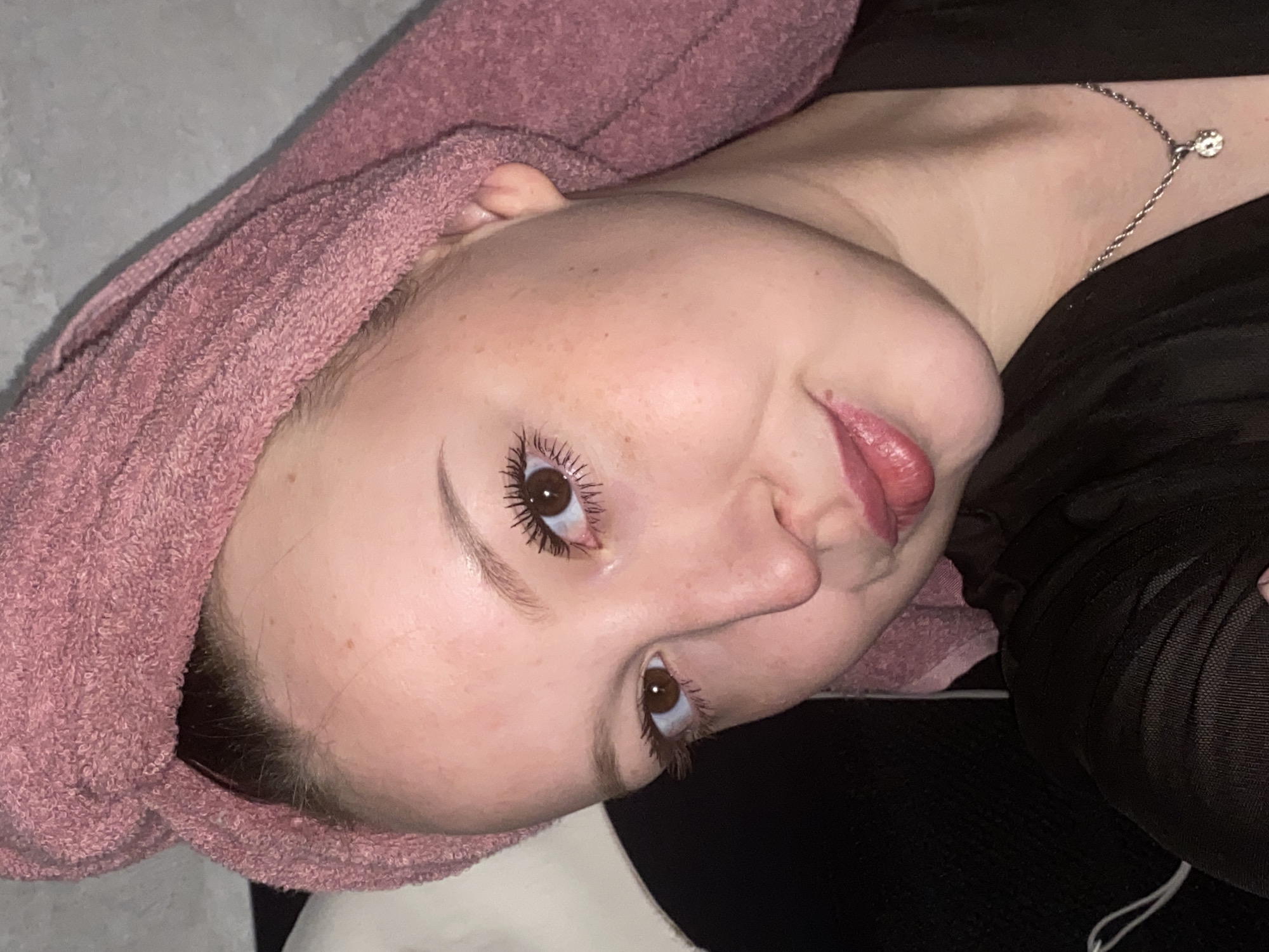 A close-up, rotated image shows a woman with a pink towel wrapped around her head. She has fair skin with freckles and brown eyes with mascara. She is wearing lipstick. A silver chain necklace is visible on her neck. She is wearing a black top. The background is a mix of gray and black. The location is not discernible from the image.