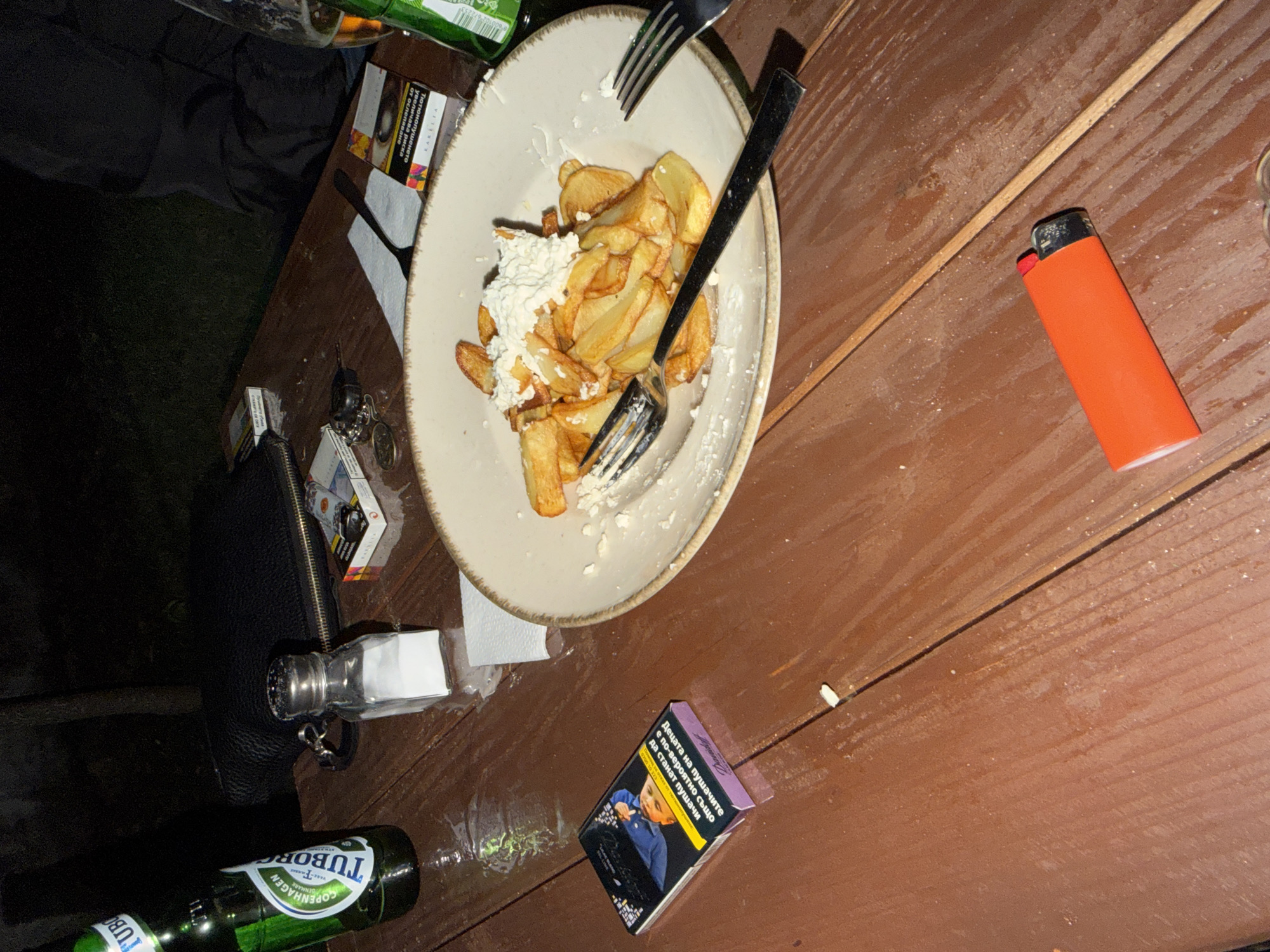 An overhead medium shot shows a wooden table with food, drinks, and smoking paraphernalia. On the left, a green bottle of Tuborg beer is partially visible. Next to it is a salt shaker filled with white salt. In the center, a plate holds a serving of french fries topped with white cheese. Two forks rest on the plate. To the left of the plate, a pack of Davidoff cigarettes with a young boy on the packaging is laid open. Several other cigarette packs and a lighter are scattered on the table. The table is wet in some areas, suggesting it has been raining or drinks have spilled. The background is dark and out of focus, indicating an outdoor setting at night.