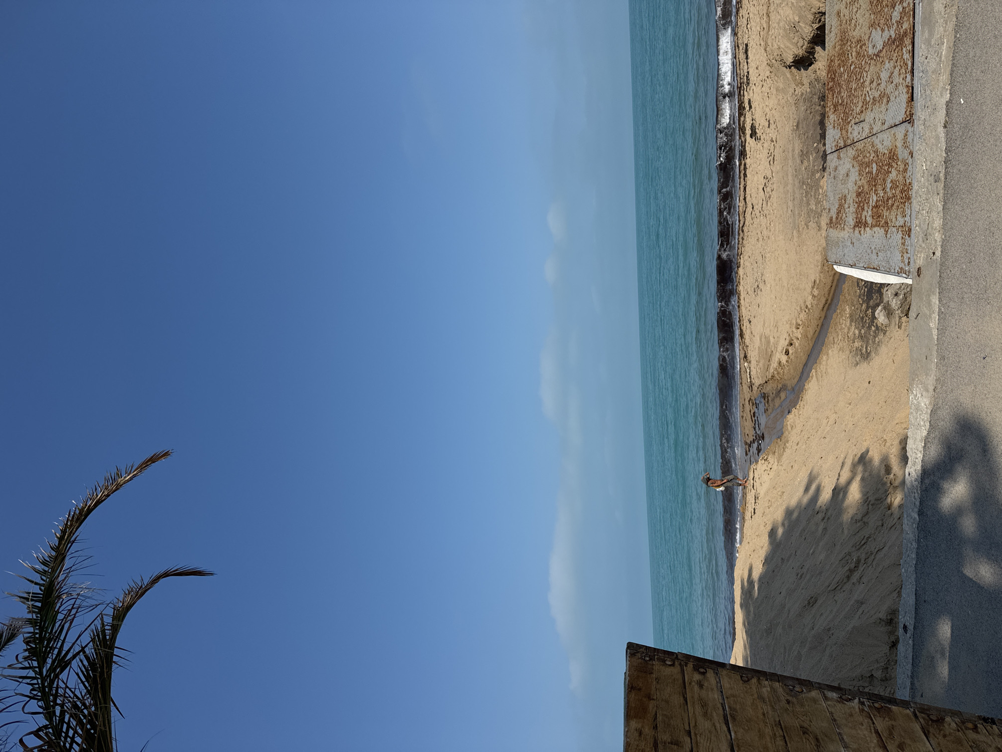 A coastal landscape is depicted, featuring a clear blue sky, calm turquoise water, and a sandy beach. The image is captured with a low horizon line, showing a vast expanse of sky.
In the foreground, on the left side, the dark green fronds of a palm tree are visible, extending upwards into the blue sky. To the right, along the shoreline, the light blue sea gently meets the tan-colored sand. Small waves are breaking at the water's edge.
On the sandy beach, towards the middle right of the frame, a lone person is standing or crouching near the water. Further to the right, on the beach itself, there is a rusty, weathered metal structure, possibly an access hatch or cover, embedded in the sand. Adjacent to the sand is a paved area, which appears to be a walkway or road. Long, dark shadows, likely cast by unseen objects or structures, stretch across the sand and the paved surface, suggesting it is either early morning or late afternoon.
In the bottom right corner, a portion of a wooden structure, possibly a fence or part of a building, is visible, partially framing the scene. The weather appears clear and sunny. There is no visible text in the image.