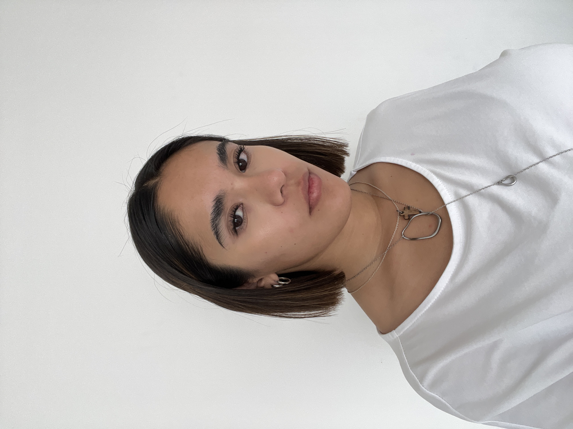 A woman with brown hair in a short bob is wearing a white t-shirt and two silver necklaces. She is looking at the camera with a neutral expression. The background is a plain white wall. The image does not appear to be in San Bernardino Tlaxcalancingo, Mexico; this information cannot be confirmed.