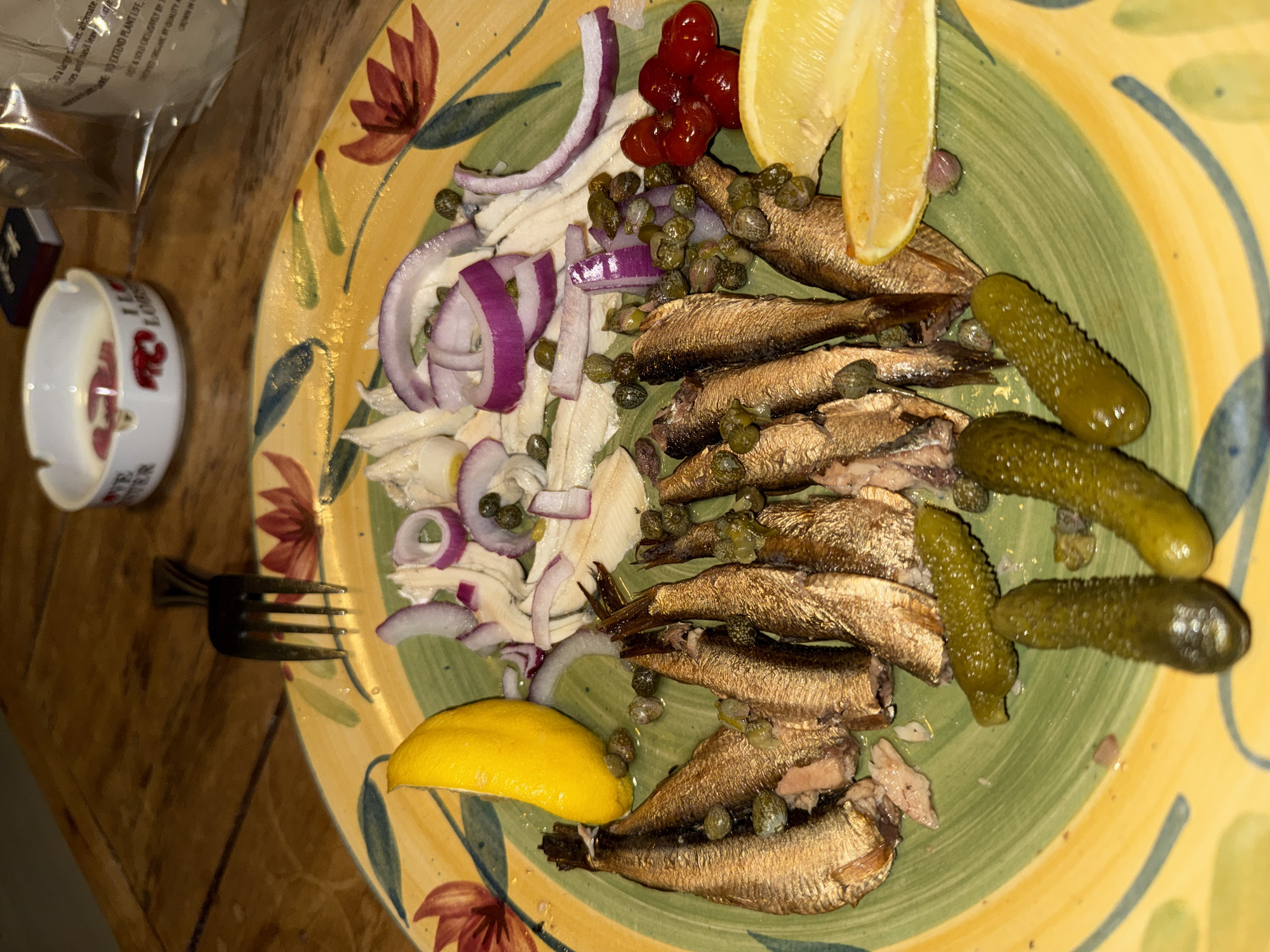 The image depicts a plate of food. The plate is ceramic, decorated with floral patterns in red, yellow, and green. Arranged on the plate are a stack of smoked sprats topped with capers, slices of red onion, pickled gherkins, two lemon wedges, and an unidentified pile of red berries. A fork lies to the left of the plate on a wooden surface. Adjacent to the fork is a white ashtray featuring a red lobster design. Behind the plate, a transparent plastic bag and a small black box are visible. The setting appears to be an indoor dining area. Since Provincetown, United States of America, is a popular fishing town known for its beaches, food and art scene, the image might have been taken at a seafood restaurant or a private residence.