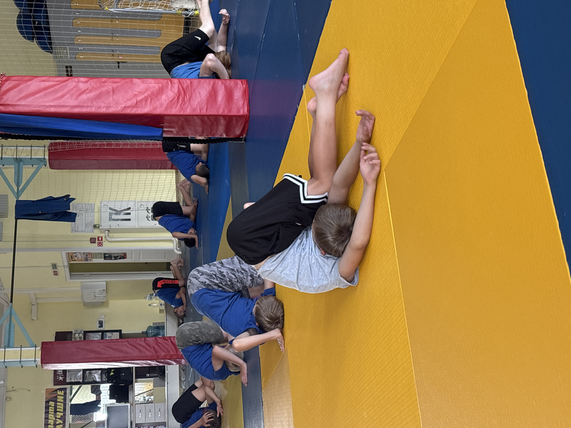 In Chaikovsky, Russia, a group of young boys are engaged in a physical training session on a martial arts mat. The scene takes place indoors, likely in a gymnasium or dojo, indicated by the presence of mats, padded pillars, and a mesh fence in the background, possibly part of a sports equipment setup. The floor is covered with blue and yellow mats. The boys, dressed in athletic wear, are performing a stretching exercise where they are on their knees, with their heads and upper bodies bent forward and down towards the mat. Their faces are mostly obscured as they are positioned with their heads down. Some boys wear blue t-shirts and black shorts, while others have camouflage patterned shorts. In the background, there are signs on the wall, including one that says "НАГМА" and another with "Детско-юношеская спортивная школа" (Children and Youth Sports School), suggesting the location is a sports facility. There are also water bottles and sports equipment visible.