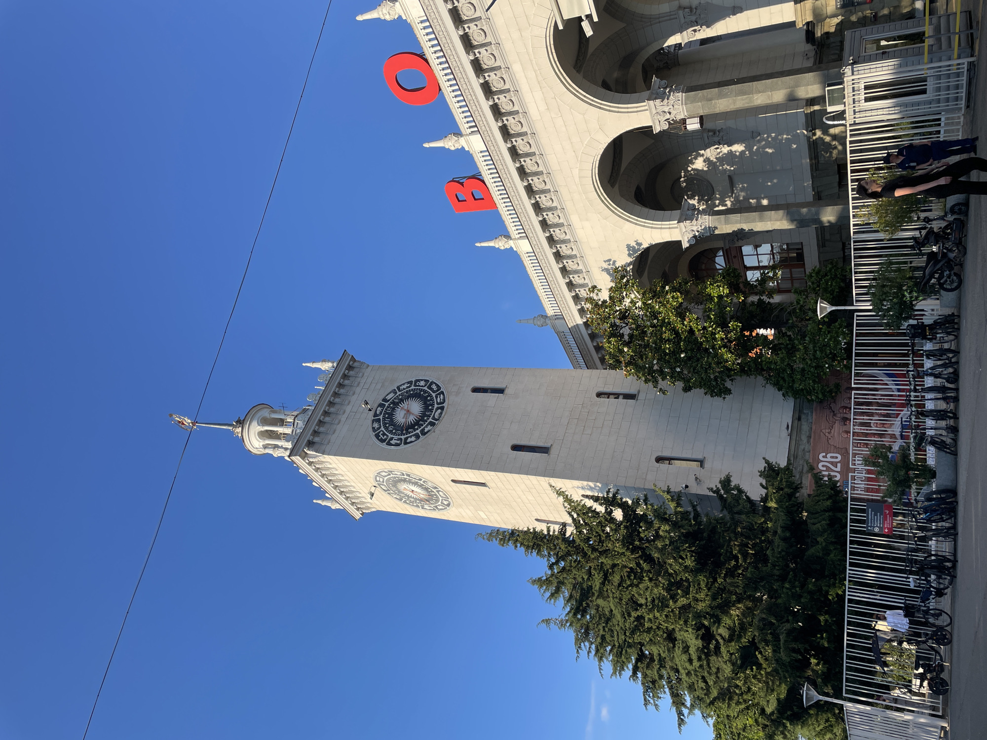 The image shows the Sochi Railway Station in Sochi, Russia. A tall, light-colored clock tower with intricate zodiac designs on its clock faces dominates the left side of the frame. Evergreen trees are visible in front of the tower. To the right, a grand building with arched windows and ornate architectural details is partially visible. Large red letters spelling "В О" (which likely form part of a larger word or acronym) are mounted on the building's facade. A white fence runs across the foreground, with several bicycles and electric scooters parked behind it. A person in a white shirt stands near the bicycles. The sky is a clear, bright blue. Shadows from the building and trees are cast across the scene, suggesting it is a sunny day.