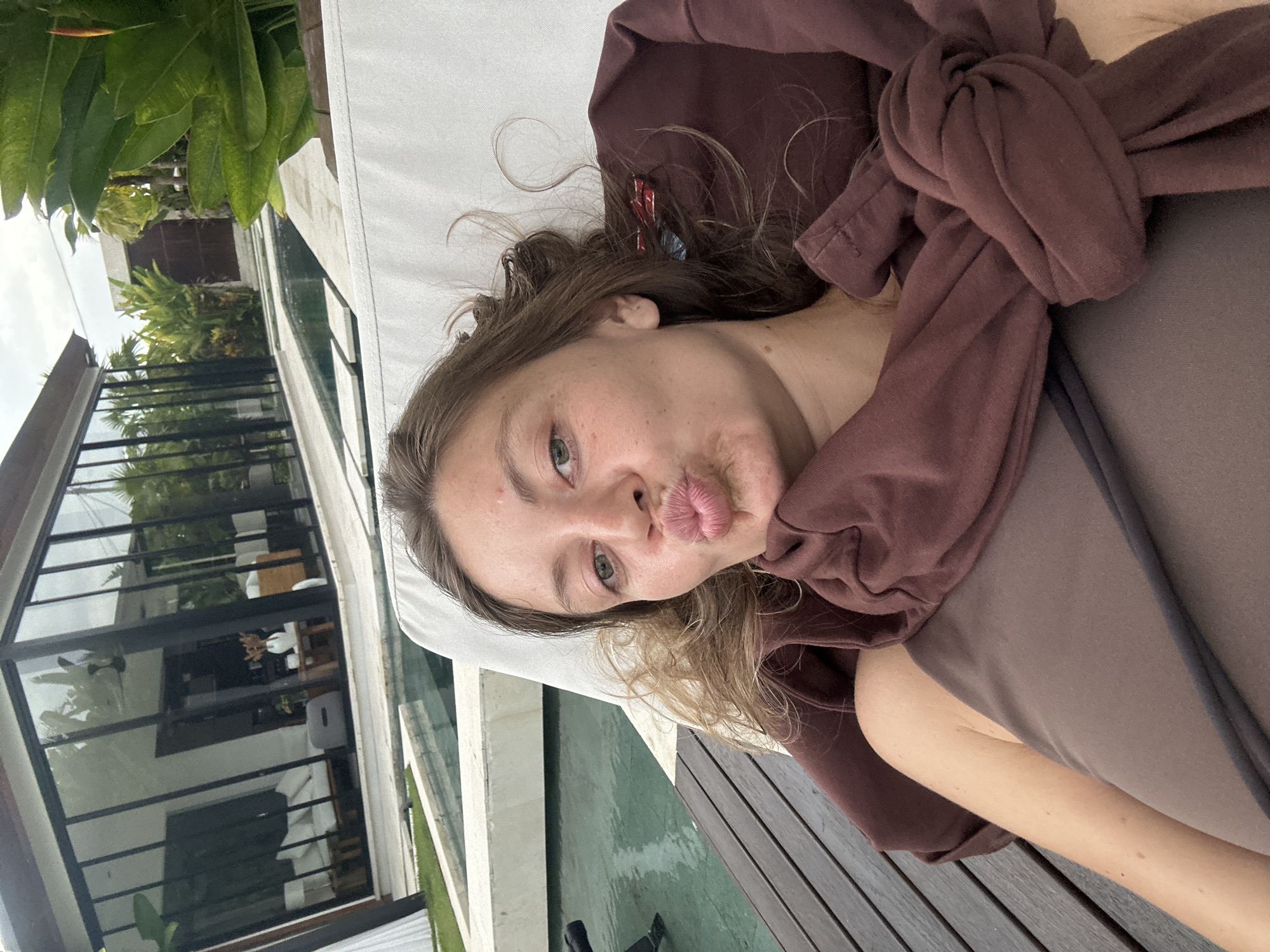 A woman with light skin, freckles, and light brown hair is lying on a daybed, making a kissing face at the camera. She is wearing a brown sweatshirt tied in a knot. The daybed has a white cushion and a gray frame with wooden slats. There is a building with large windows behind the daybed, with green plants visible through the windows. A pool of water is between the daybed and the building. The location appears to be in Ubud District, Indonesia, based on the architecture and the surrounding vegetation.