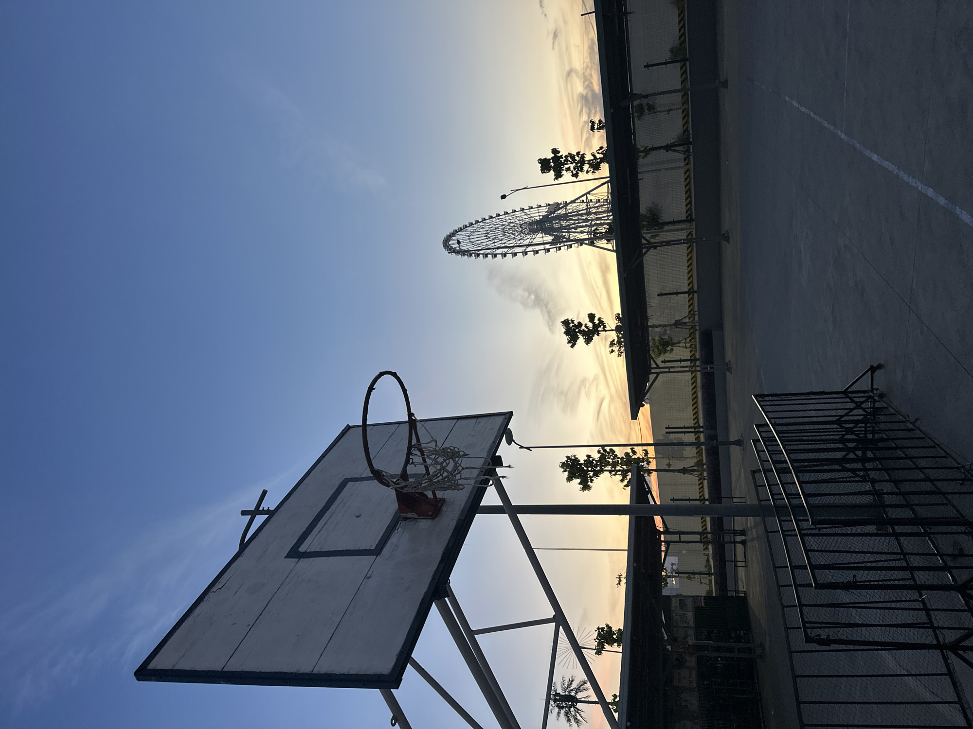 The image captures an outdoor basketball court in Da Nang, Vietnam, at dusk. A weathered basketball hoop with a torn net is prominent in the foreground, silhouetted against a clear blue sky with wispy clouds. In the background, a large Ferris wheel stands tall, partially obscured by the sunset's warm hues. The court itself is made of concrete, with faint white lines marking its boundaries. A black metal fence encloses part of the court, and several small trees and covered structures are visible along the perimeter. The overall atmosphere is serene, with the fading daylight casting long shadows.