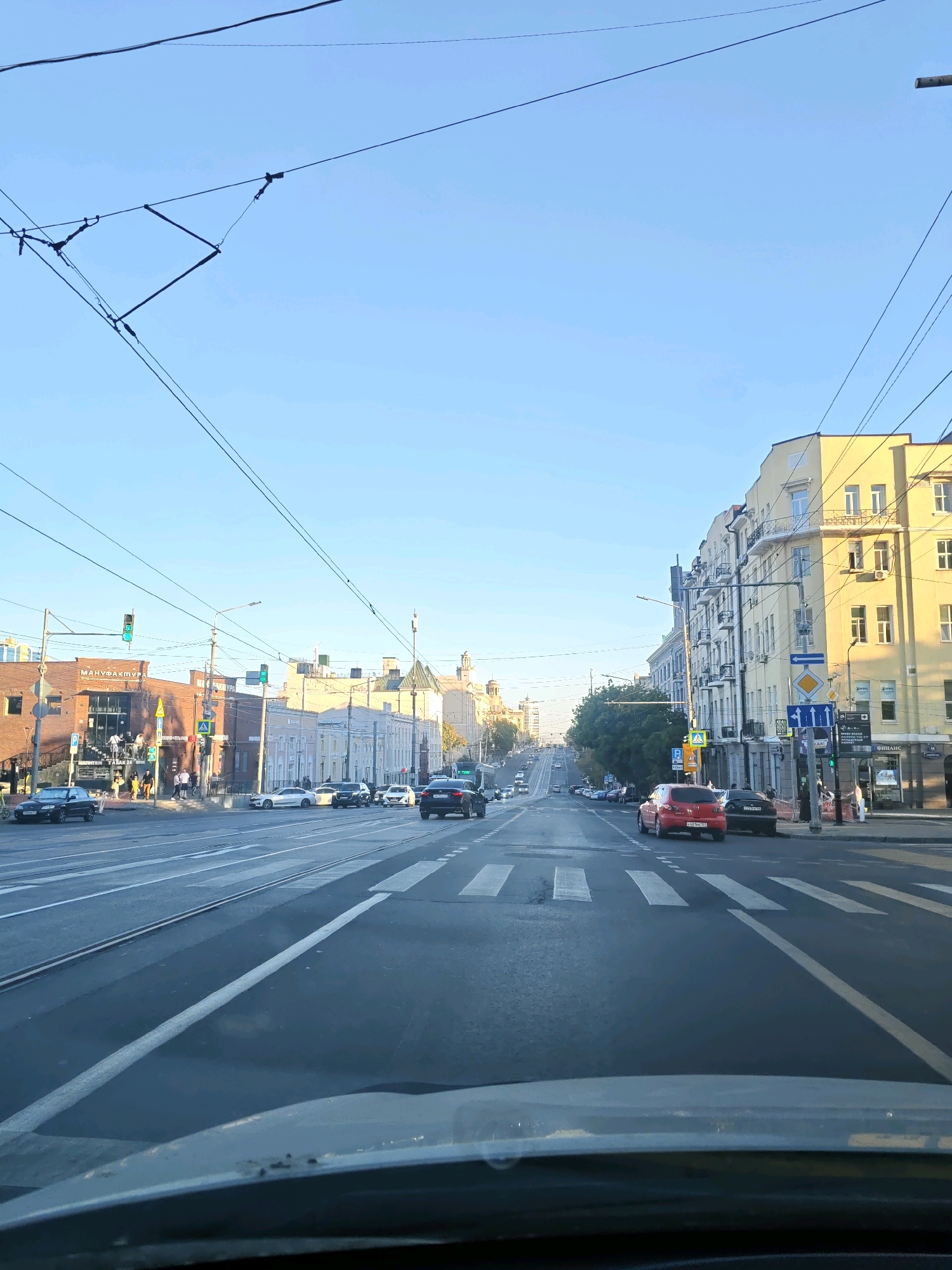 The image depicts a street scene in Rostov-on-Don, Russia, likely on a clear day with the sun casting long shadows. The perspective is from inside a vehicle, looking forward down a wide, multi-lane street lined with buildings and trees. Overhead, a network of power lines and trolley wires crisscrosses the sky.
In the foreground, the dashboard of the vehicle is visible. The street itself has white lane markings and pedestrian crossings. Several cars are driving on the street, including a black sedan on the left, a white sedan and a black SUV in the middle lanes, and a red hatchback and a black sedan on the right.
On the left side of the street, a brick building with the sign "МАНУФАКТУРА" (Manufactura) is prominent, with several people visible on its steps and sidewalk. Adjacent to it are other buildings with storefronts and windows. Traffic lights are visible, some showing green.
On the right side of the street, a tall, light-yellow apartment building with multiple windows and balconies dominates the view. Smaller buildings and storefronts are also present, including one with a sign that says "ФИНАНС" (Finance). Street signs and traffic signals are also visible.
In the distance, the street appears to curve and ascend, with more buildings and trees receding into the horizon. The sky is a clear, pale blue. The overall impression is that of an active urban environment with ongoing traffic and pedestrian activity.