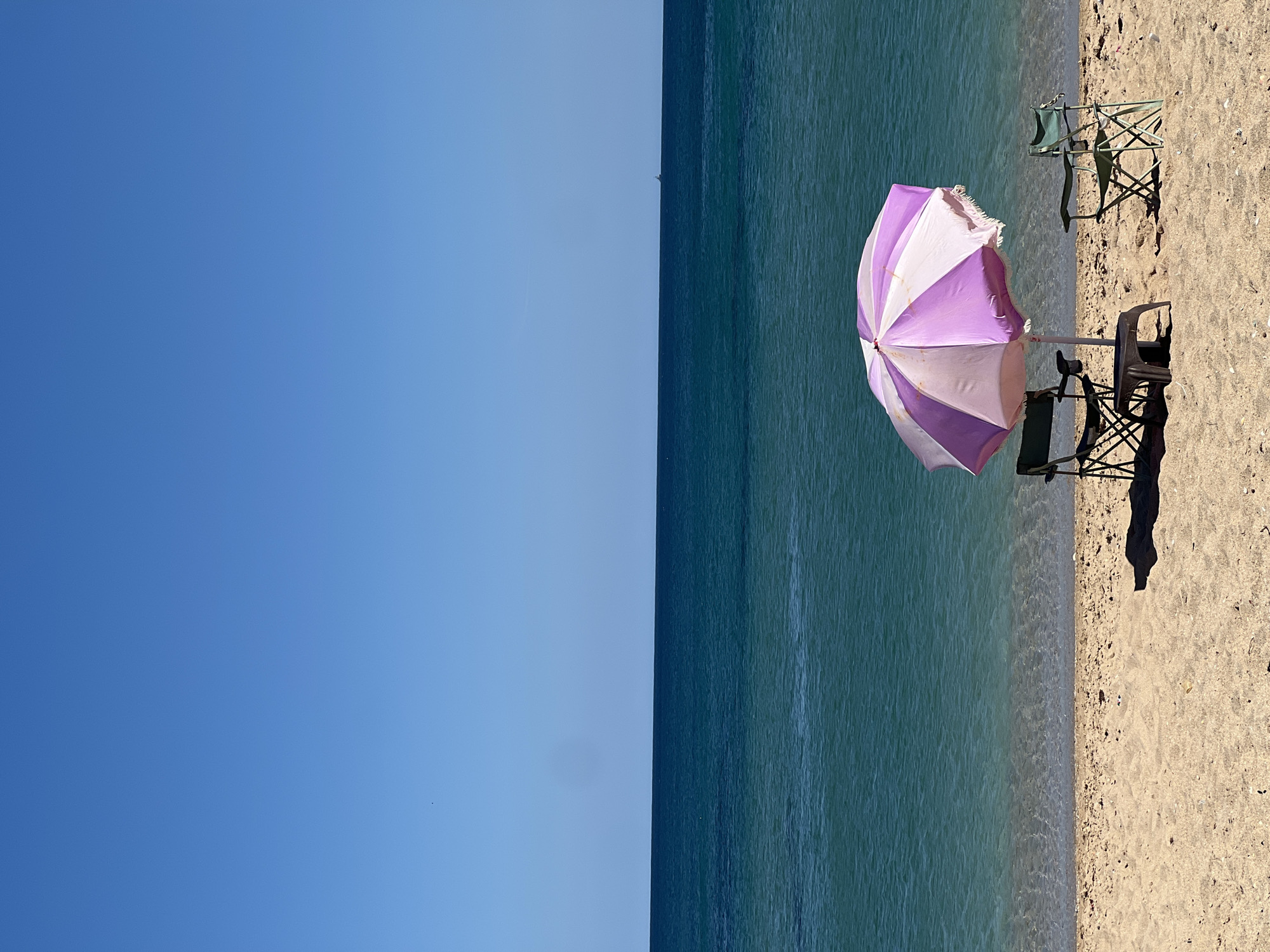 The image depicts a beach scene in M'Diq, Morocco. The sky is clear blue, transitioning to a turquoise sea. The right side of the image shows a sandy beach with a striped pink and white beach umbrella, casting a shadow. Two chairs are visible near the umbrella, and two more chairs are positioned further away on the sand. The image is oriented vertically, with the horizon line running down the center.