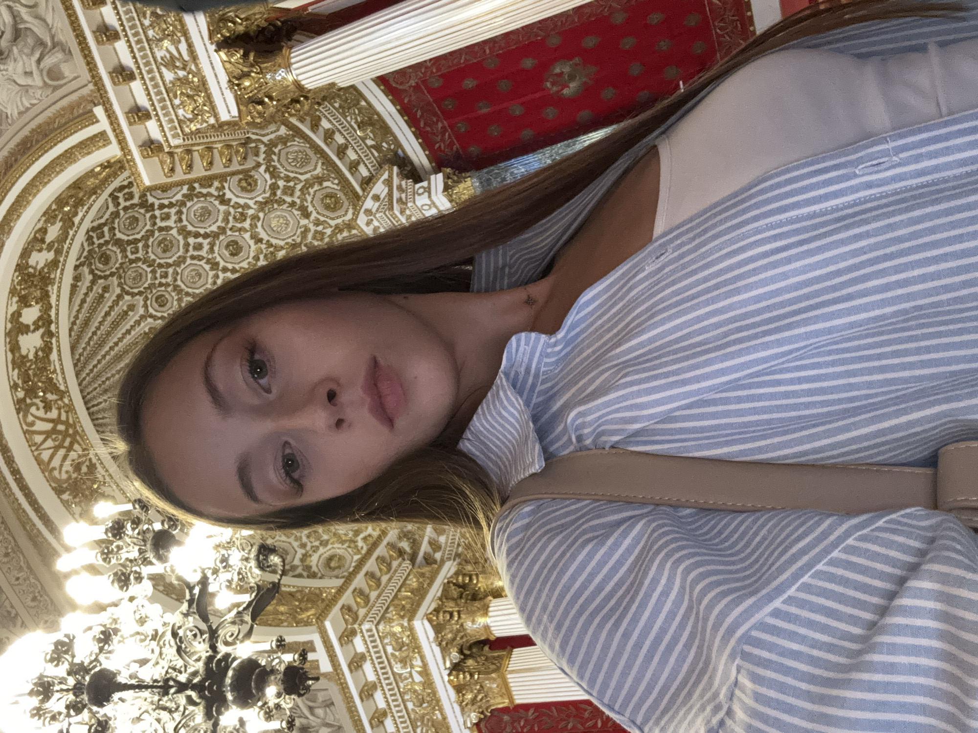 The media file is a selfie taken by a young woman in an ornate, indoor setting. She is wearing a light-colored top and a blue and white striped button-down shirt, with a beige strap across her shoulder, possibly from a bag. Her long, brown hair is down, framing her face. The background is richly decorated with gold accents, intricate patterns, and a red ceiling with gold detailing, suggesting a historical or palatial environment. A large, dark chandelier is visible in the lower part of the frame. The lighting appears to be artificial, likely from the chandelier and ambient room lighting. The woman's expression is neutral, and she is looking directly at the camera. The image is oriented vertically, with the top of her head towards the bottom of the frame. No visible text is present.