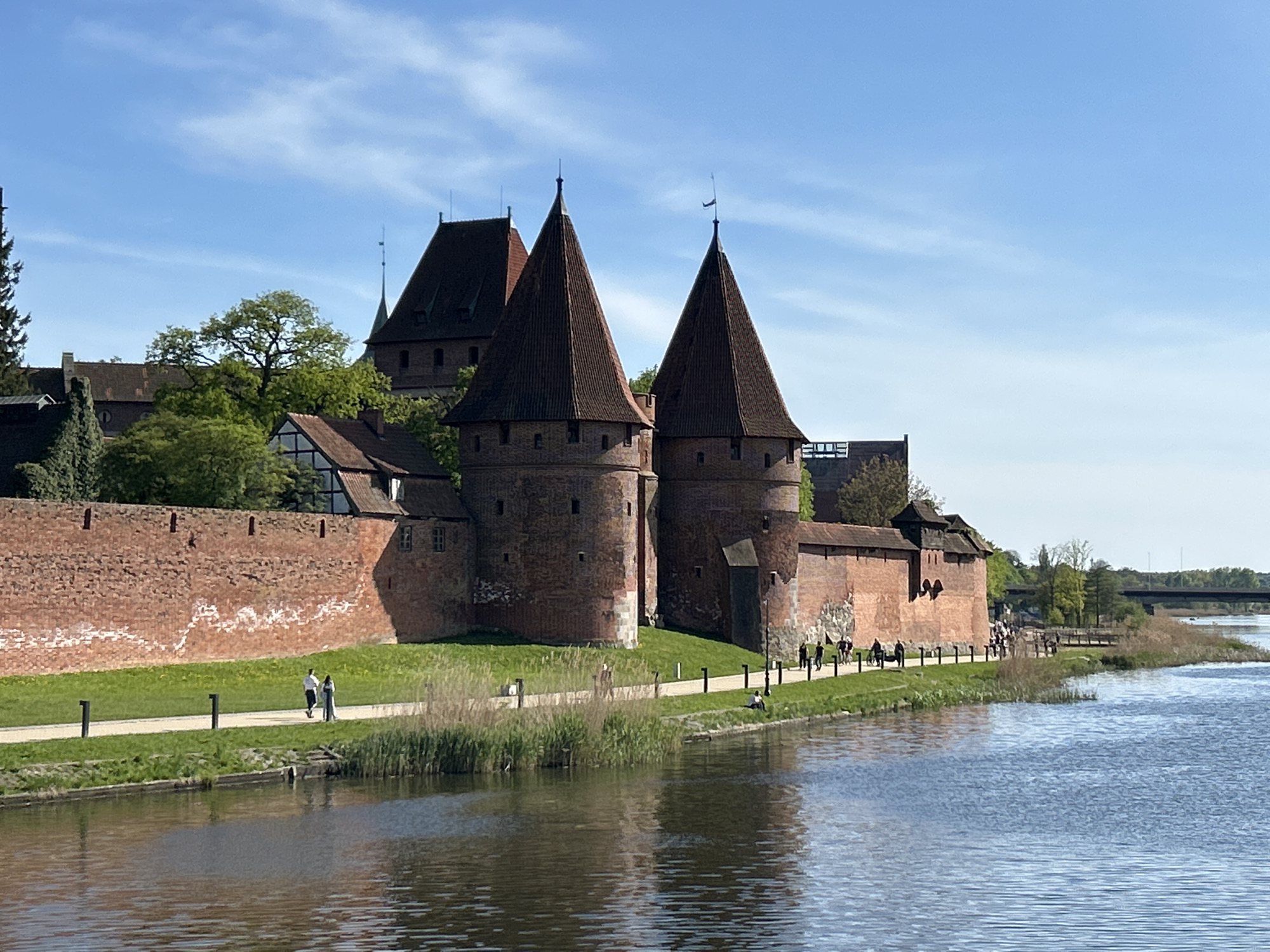 The image shows Malbork Castle in Malbork, Poland, viewed from across the Nogat River. The castle's brick walls, towers with red-tiled roofs, and other buildings are prominent. A paved walkway runs along the riverbank with grass on either side. Several people are walking on the path. The water reflects the castle and the sky, which is blue with some clouds. Reeds grow along the edge of the river. In the background, trees and a bridge are visible.