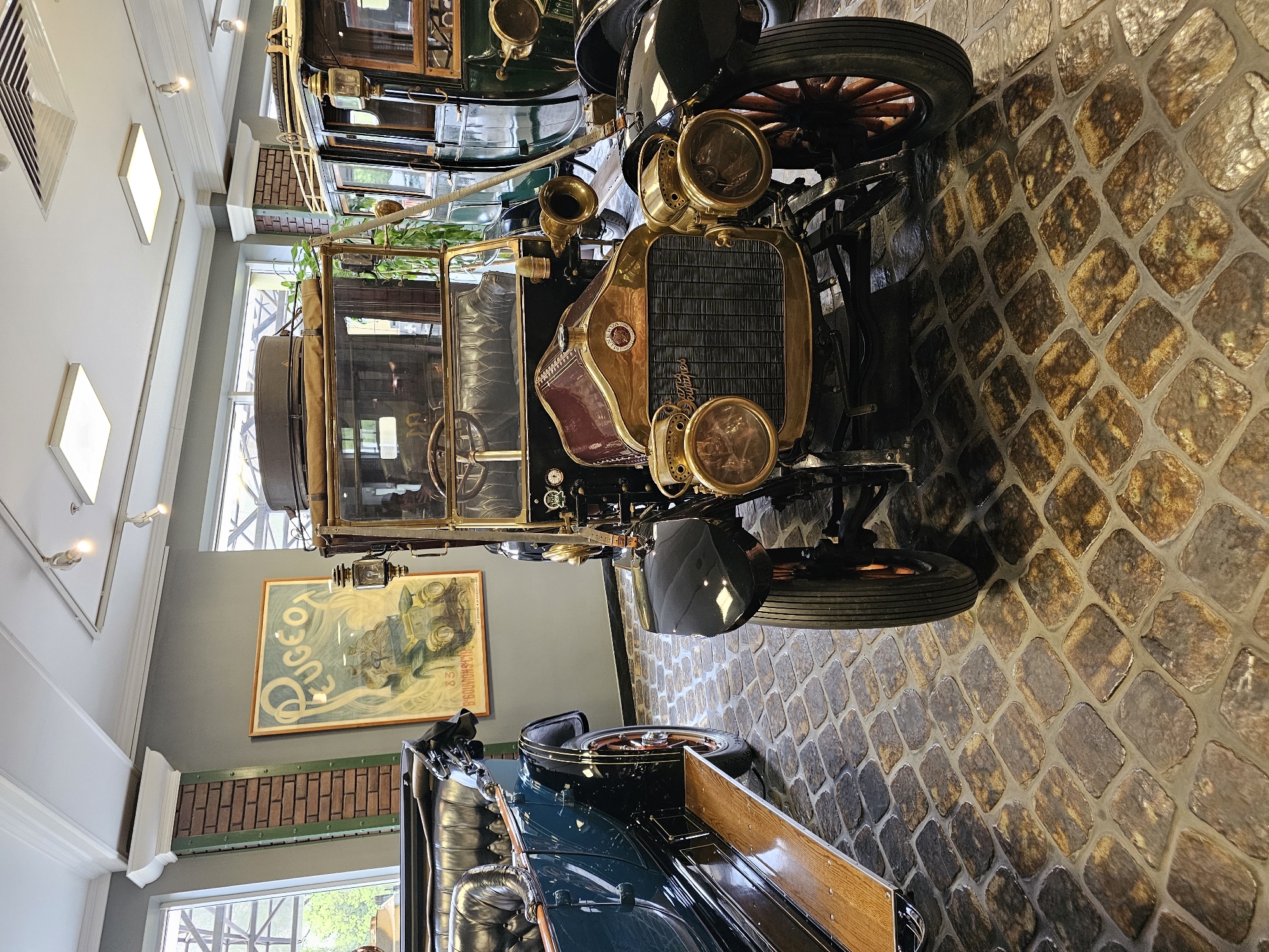 The image shows an interior view of a museum or exhibition hall, likely featuring vintage automobiles. The location is possibly Архангельское, Russia, inferred from the presence of vintage cars, which is consistent with museum or historical displays.
The foreground is dominated by an antique car with a black chassis, brass or bronze-colored accents, and large, classic headlights. The car has a radiator with a black mesh grille. Its tires are thin and likely made of solid rubber.
In the background, other vintage cars are partially visible. There's a large poster of a Peugeot car on a wall, framed and hung above a faux brick wall.
The floor is paved with roughly textured stones, creating a vintage ambiance. The lighting is provided by modern fixtures on the ceiling, creating an artificial illumination environment. The setting suggests a controlled, museum-like environment for displaying and preserving these historical vehicles. There are no people visible in the frame.
