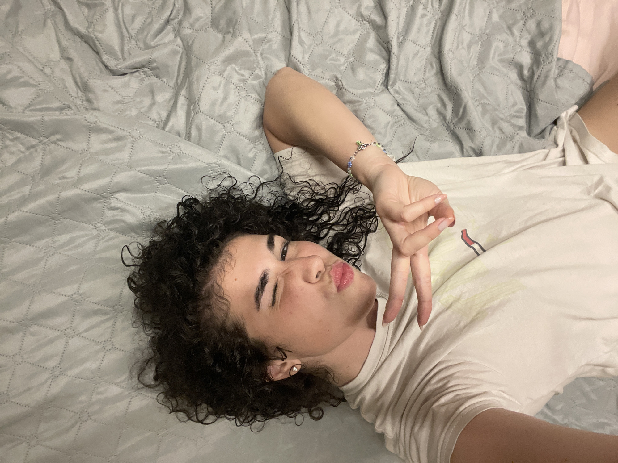 A person with curly dark hair and light skin is lying on a gray quilted surface, likely a bed or blanket. They are wearing a white t-shirt and have their right eye closed in a wink, with their lips pursed. Their left hand is raised, forming a peace sign, with painted nails. A beaded bracelet is visible on their right wrist. The background is a gray, quilted surface. The scene does not provide any information to determine a location in Sofia, Bulgaria.