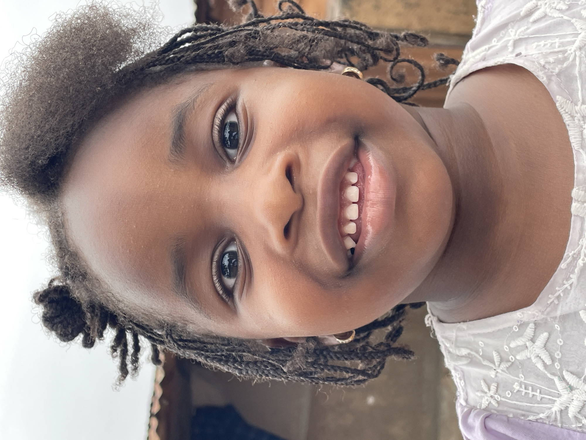 A close-up, slightly tilted image shows a young girl with brown skin smiling at the camera. She has braided hair with a section of her natural hair at the top of her head. She is wearing a white lace top with a hint of lavender underneath. Gold earrings are visible in her ears. While there is some background visible, there are no specific details to indicate the location is Jos, Nigeria.