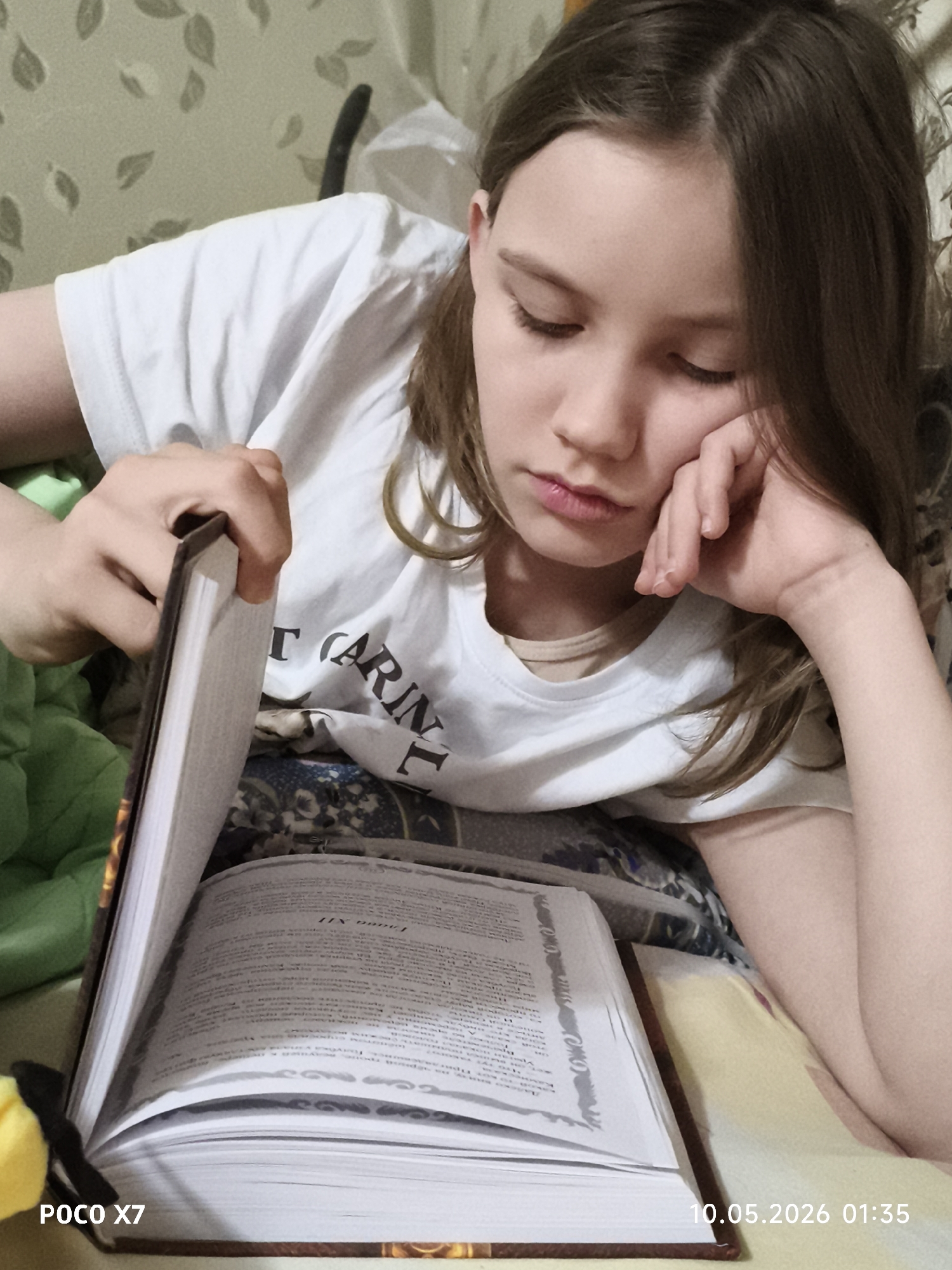 A young person is lying down while reading a book. They are wearing a white shirt and the book is open in front of them. The text in the book is in a different language. The location of the city is Unknown, Unknown. A green blanket is visible in the background. The time stamp on the image is 10.05.2026 01:35.