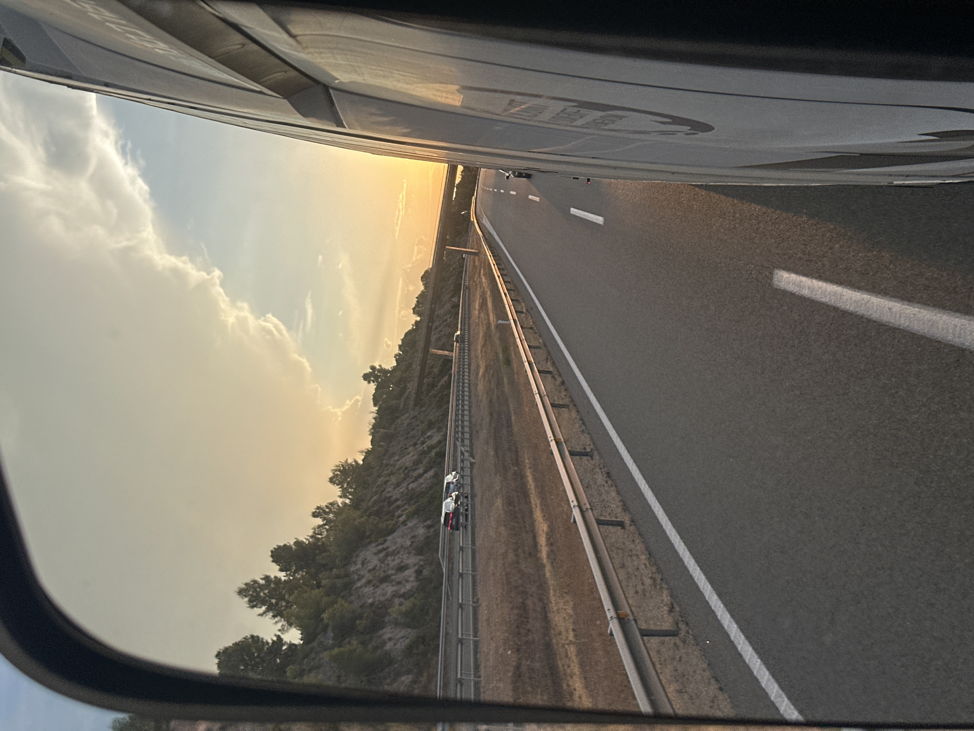 The image, taken from the perspective of a vehicle, captures a highway scene at sunset. A golden sunset illuminates the sky, casting a warm glow on the clouds. The highway stretches into the distance, with a line of cars visible ahead. The roadside is lined with dry vegetation and a protective barrier. On the left side of the frame, a steep, rocky incline covered in trees rises from the highway. The right side of the image is dominated by the side of a vehicle, possibly a truck or van, with visible branding. The scene appears to be on a highway outside the city of Bràfim, Spain, judging by the characteristic dry, hilly landscape and the presence of a highway.