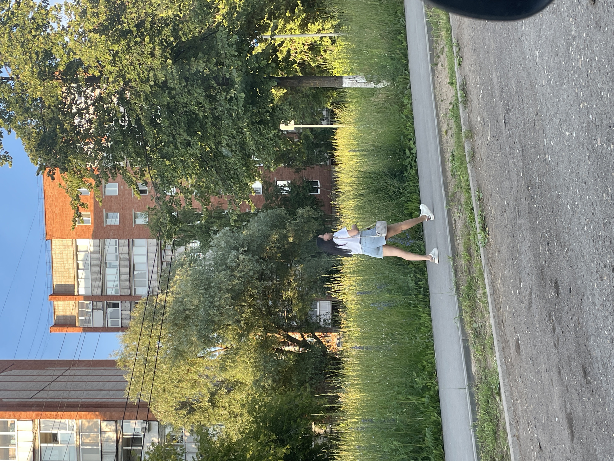 In Votkinsk, Russia, a young woman with dark hair walks along a paved sidewalk. She is wearing a white t-shirt and denim shorts, with a small, light-colored purse slung over her shoulder. White sneakers adorn her feet. The sidewalk runs alongside a dense area of tall green grass, interspersed with purple wildflowers. Behind the grassy area, a line of trees with lush green foliage can be seen. In the background, two multi-story brick apartment buildings with numerous windows stand under a clear blue sky. Power lines are visible crisscrossing the sky between the buildings and the trees. The ground in the foreground appears to be a dirt road or unpaved area, with scattered stones and patches of dry earth. The scene is illuminated by natural sunlight, suggesting it is daytime.