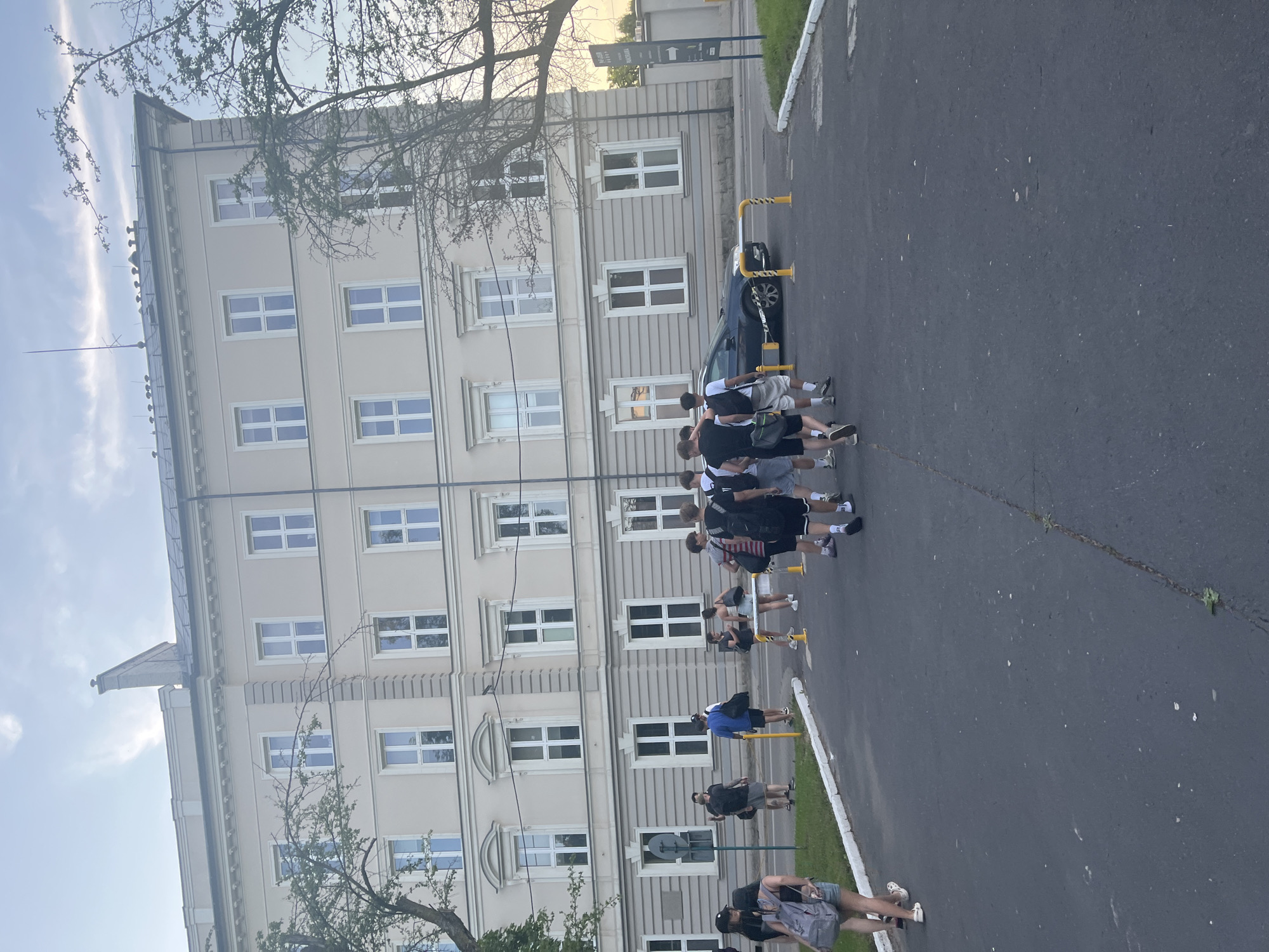 The image captures a street-level view of a multi-story building with a light-colored facade and numerous evenly spaced windows. In front of the building, a group of approximately ten individuals, predominantly young adults, are walking on an asphalt surface. The group appears to be moving in the same direction, away from the camera. Some individuals are carrying backpacks, and their attire suggests casual wear. A dark-colored sedan is parked on the right side of the road, partially obstructed by a yellow and black barrier. The environment includes trees with sparse foliage and a cloudy sky. The scene seems to be set in an urban or suburban area, possibly near an educational institution given the building's architecture and the presence of young people.