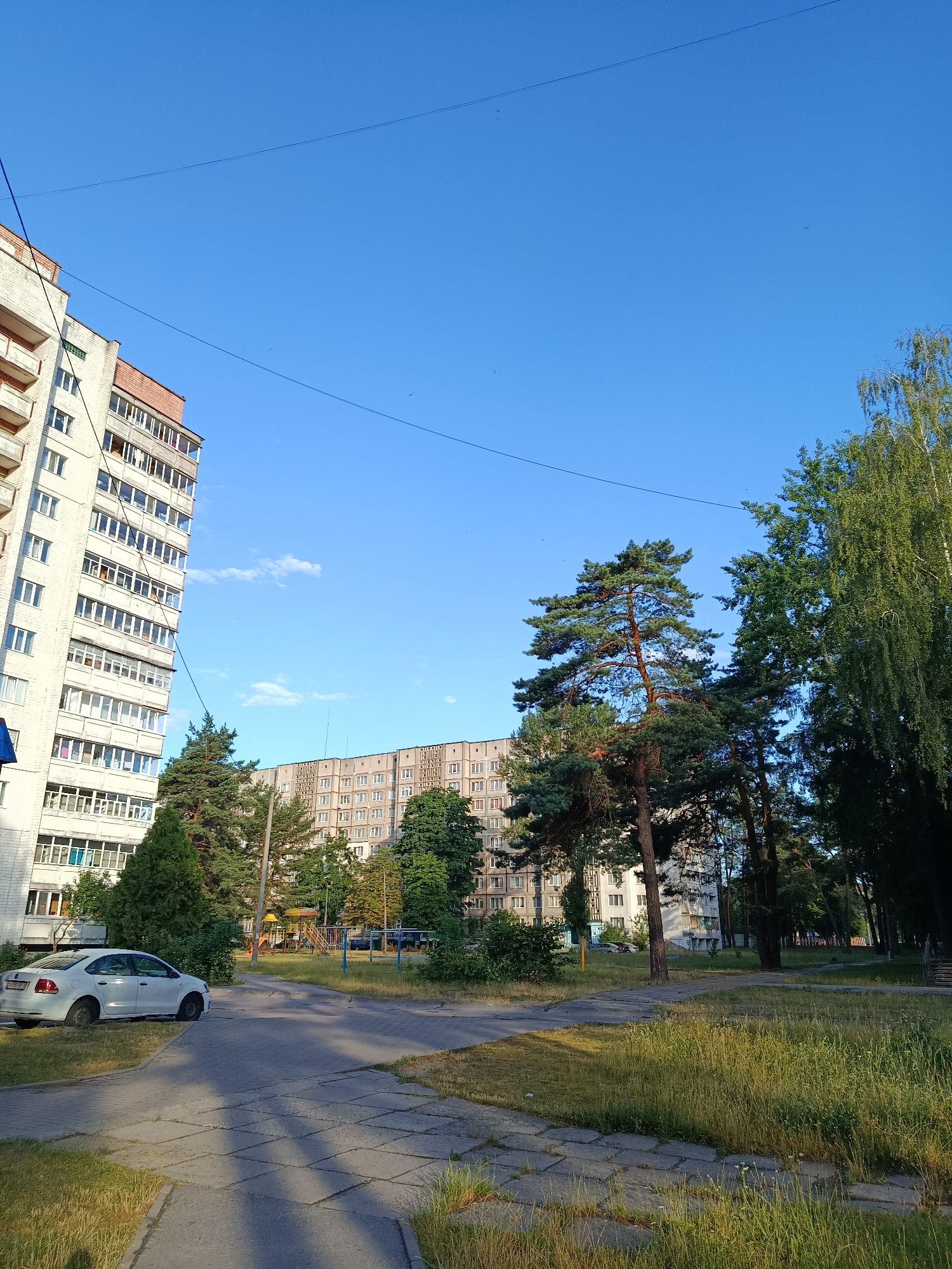The image depicts a residential courtyard in the city of Svietlahorsk, Belarus, on a clear day with a bright blue sky. On the left side of the frame, a multi-story apartment building with a light-colored facade and red brick accents dominates the scene. The building features numerous windows and balconies. In front of this building, a white sedan is parked on a paved area.
To the right of the apartment building, a playground with colorful structures is visible amongst trees and bushes. Further into the courtyard, another long, light-brown apartment building is seen, characterized by its repetitive window pattern. Several tall trees, including pines and birches, are scattered throughout the courtyard, casting shadows on the paved walkways and grassy areas. The ground is a mix of concrete pathways and overgrown grass. The overall impression is of a typical Soviet-era housing complex, with well-established greenery.