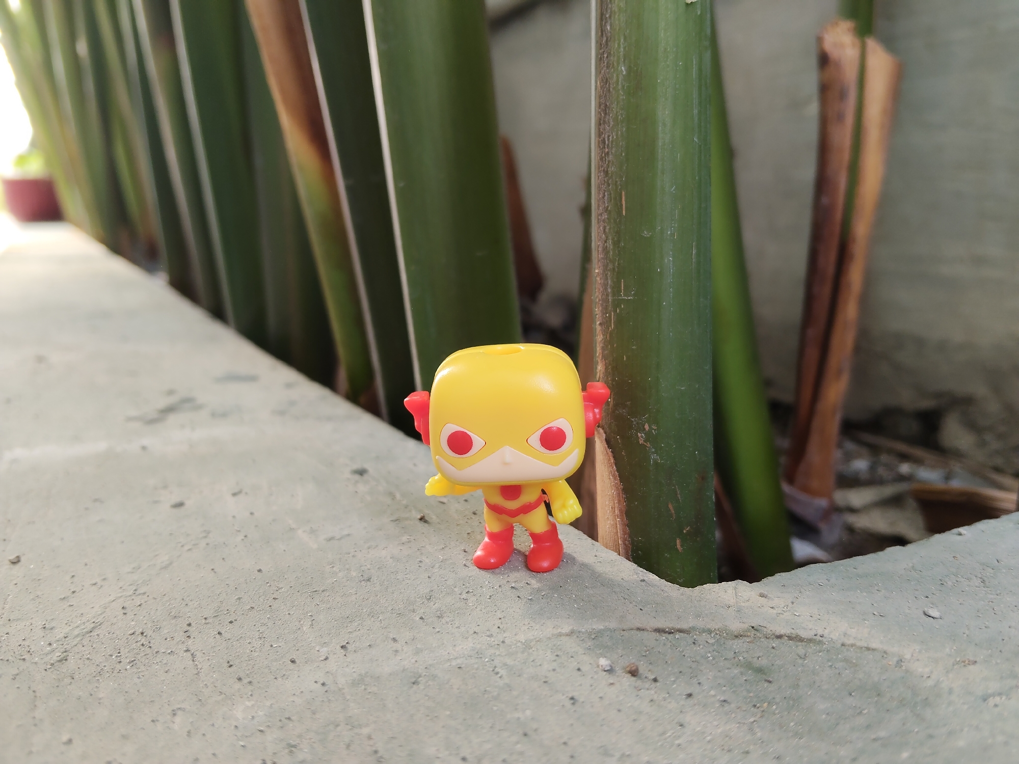 A small yellow toy figurine resembling the Flash superhero stands on a grey concrete surface. The figurine has red accents on its mask, boots, and lightning bolt ear accessories. Behind the toy, a row of tall, green, bamboo-like stalks runs diagonally across the frame, with some appearing drier and browner. The background is blurred, suggesting a shallow depth of field, and hints at an outdoor urban environment, possibly a garden or pathway in Pemalang, Indonesia. The concrete surface is textured with small pebbles and appears weathered.