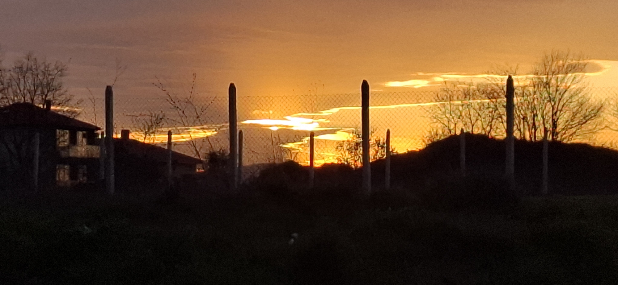 A long shot photograph was taken in Unknown, Unknown, showing a sunset with the sun low on the horizon. Silhouetted structures and objects are in the foreground. A two-story house with visible windows and a red roof can be seen on the left of the image. A series of tall, vertical posts connected by a mesh fence run across the middle of the frame. Behind the fence, there is a tree without leaves on the right side. The sky is an orange hue, suggesting either a sunrise or sunset. The ground appears dark and unclear due to the low light conditions.