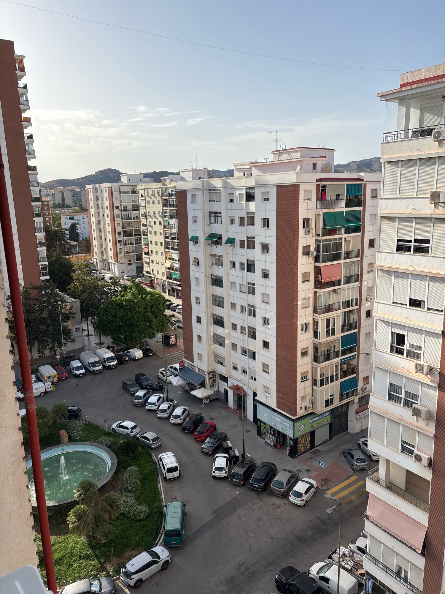 The image shows an aerial view of a street in Malaga, Spain, with several apartment buildings, parked cars, and a small park with a fountain. The buildings are multi-story residential blocks, typical of urban Spanish architecture. The street appears to be a residential area, with cars parked on both sides and in a central courtyard.
In the foreground, a circular fountain with water jets is visible within a green area. Several palm trees and manicured bushes surround the fountain. Several cars are parked around this green space, including white vans, a green van, and various passenger cars.
Further into the scene, a row of apartment buildings dominates the view. Some of these buildings have balconies with awnings and air conditioning units. Several shops are visible on the ground floor of one of the buildings, including a minimarket. A pedestrian crossing is marked on the road. The sky is clear with some scattered clouds. The overall impression is of a typical urban environment in a Mediterranean city.