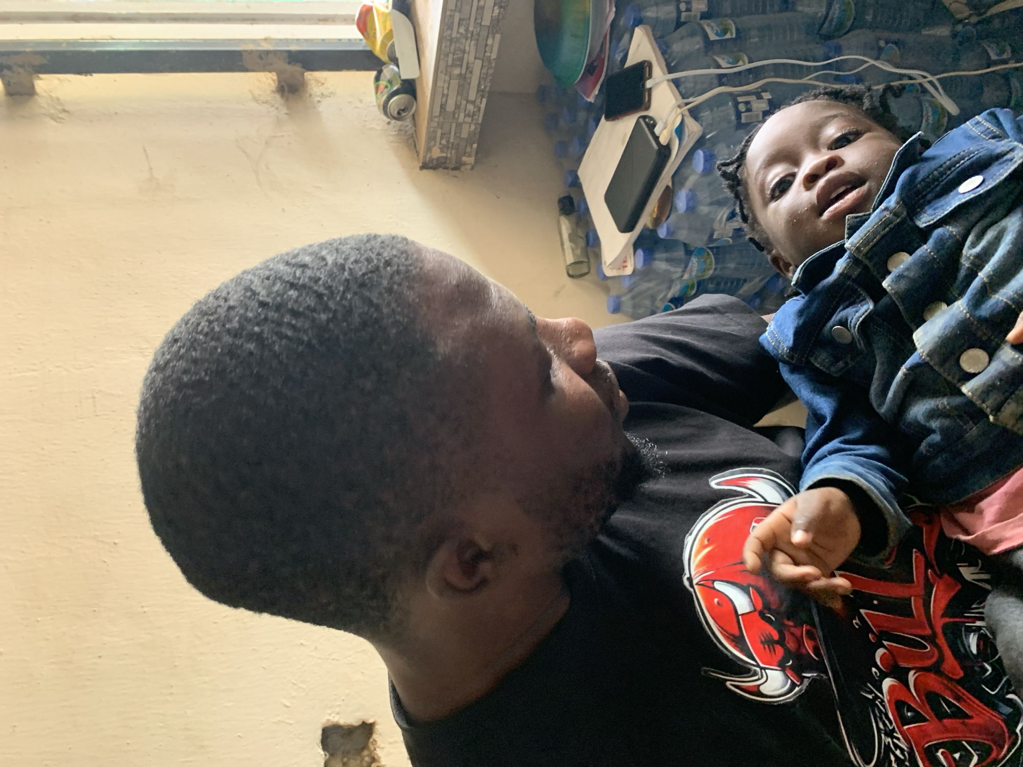 An adult male holds a young child within an indoor setting in New Stadium, Nigeria. The male, of African descent, has short, dark, wavy hair and a beard, and wears a black t-shirt featuring a red and white Chicago Bulls logo. He is positioned in the left foreground, looking down towards the child with a focused gaze. The child, also of African descent, has dark skin and hair styled in small braids or twists, and wears a blue denim jacket with silver buttons. The child lies partially on their back, facing towards the viewer with an open mouth and raised right hand.
The background reveals a light-colored, textured wall with some visible damage or wear on the left. To the right, a dark vertical frame suggests a window or opening. Below this, a tiled ledge holds two aluminum cans (one black and silver, one yellow and red) and a small white object. Beneath the ledge and behind the child, numerous transparent plastic water bottles with blue caps are stacked. Resting on top of these bottles are a white notebook, a black power bank, and a black smartphone charging via a white cable. A green plastic bowl is also visible among these items. The overall scene suggests a functional or domestic environment.