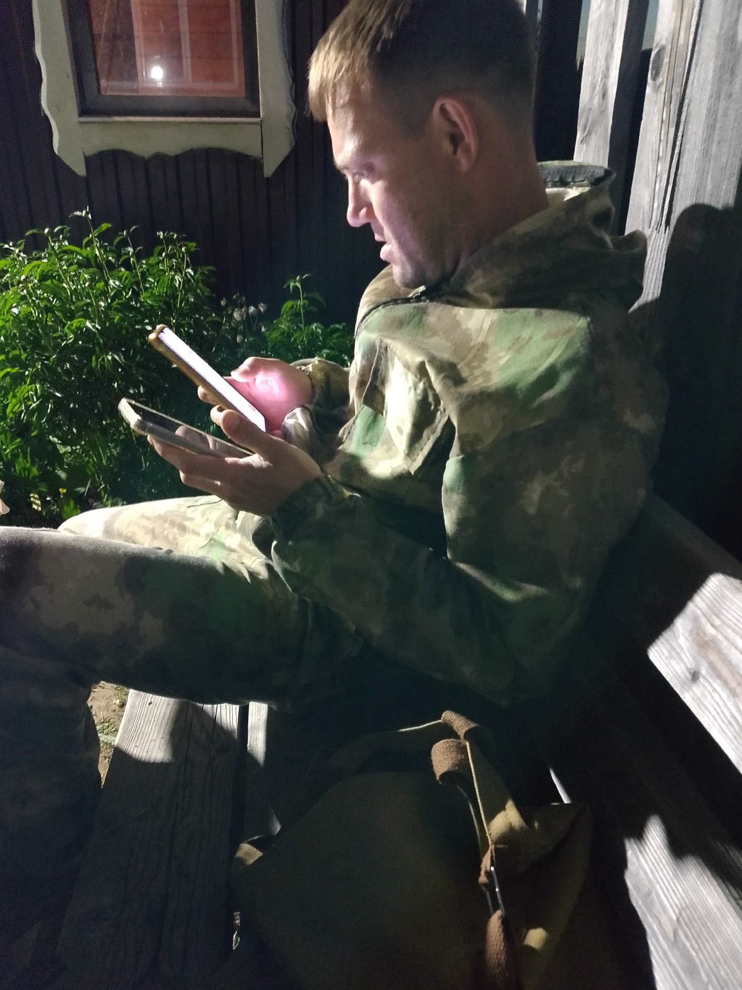 A man in a camouflage jacket and pants is seated on a wooden bench outdoors at night. He is holding two smartphones, one in each hand, and looking at them. A dark green bush is behind him on the left. A dark building is behind the bush, with a lit window visible. A canvas bag is at his feet on the bench. The location is potentially Прикамский, Russia.