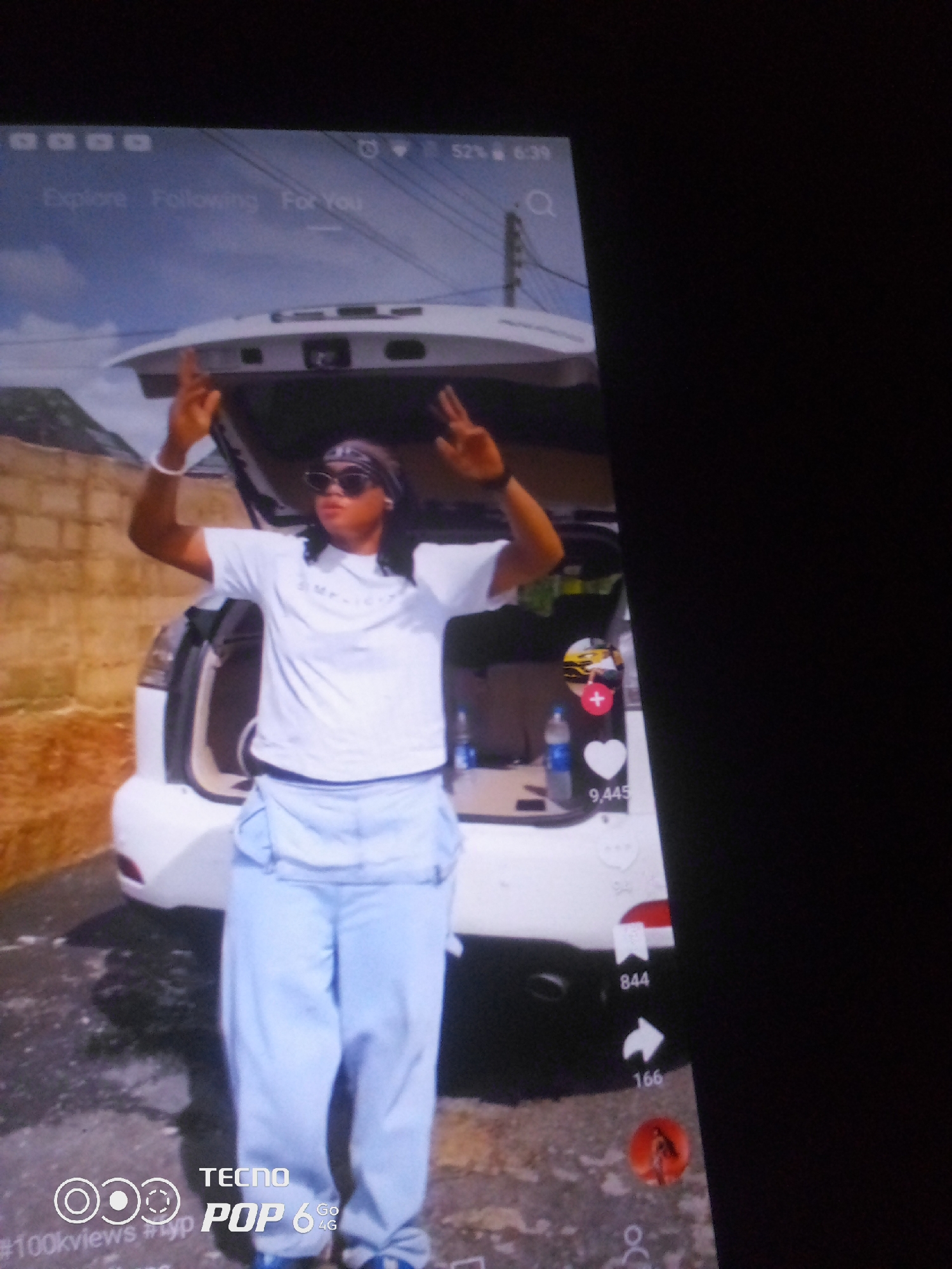 This media file captures a person standing in front of the open trunk of a white car. The person is wearing a white t-shirt, light blue overalls, a bandana, and sunglasses. They have their hands raised with fingers pointed upwards. Behind the car, a brick wall and some power lines are visible. The scene appears to be outdoors, possibly in a driveway or open area, with sunlight suggesting it's daytime.
The image also shows a digital interface overlaid on the scene, likely from a mobile device. This interface includes navigation tabs labeled "Explore," "Following," and "For You," along with a search icon. There are also social media engagement icons and numbers, such as a heart with "9,445," a bookmark with "844," and an arrow with "166." Text at the bottom indicates "TECNO POP 6 4G" and "#100kviews #Lyp." The overall impression is a candid shot of a person, potentially capturing a moment related to social media content creation or viewing, set in Howayi, Nigeria.