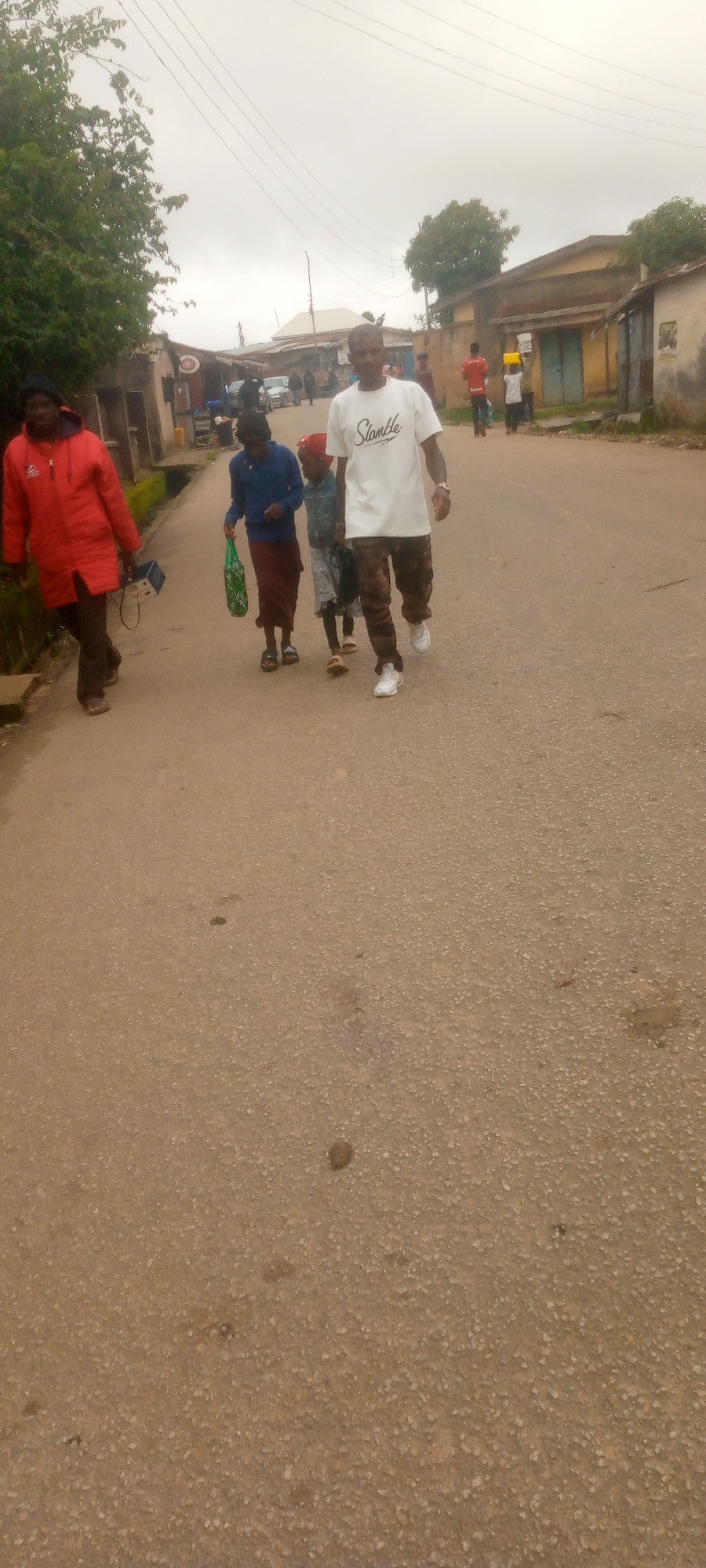 In a street scene in Unknown, Unknown, a group of people are walking on a paved road. A person in a red jacket is walking on the left side of the road, carrying a device. Three people are walking together in the center of the road: an adult wearing a blue top and a long skirt, a child wearing a red hat and patterned shorts, and another child in a patterned dress. To the right of them, a man in a white t-shirt with "Slamble" written on it and patterned pants is walking. In the background, there are buildings lining the street, some with awnings, and a few vehicles are visible. The sky is overcast.