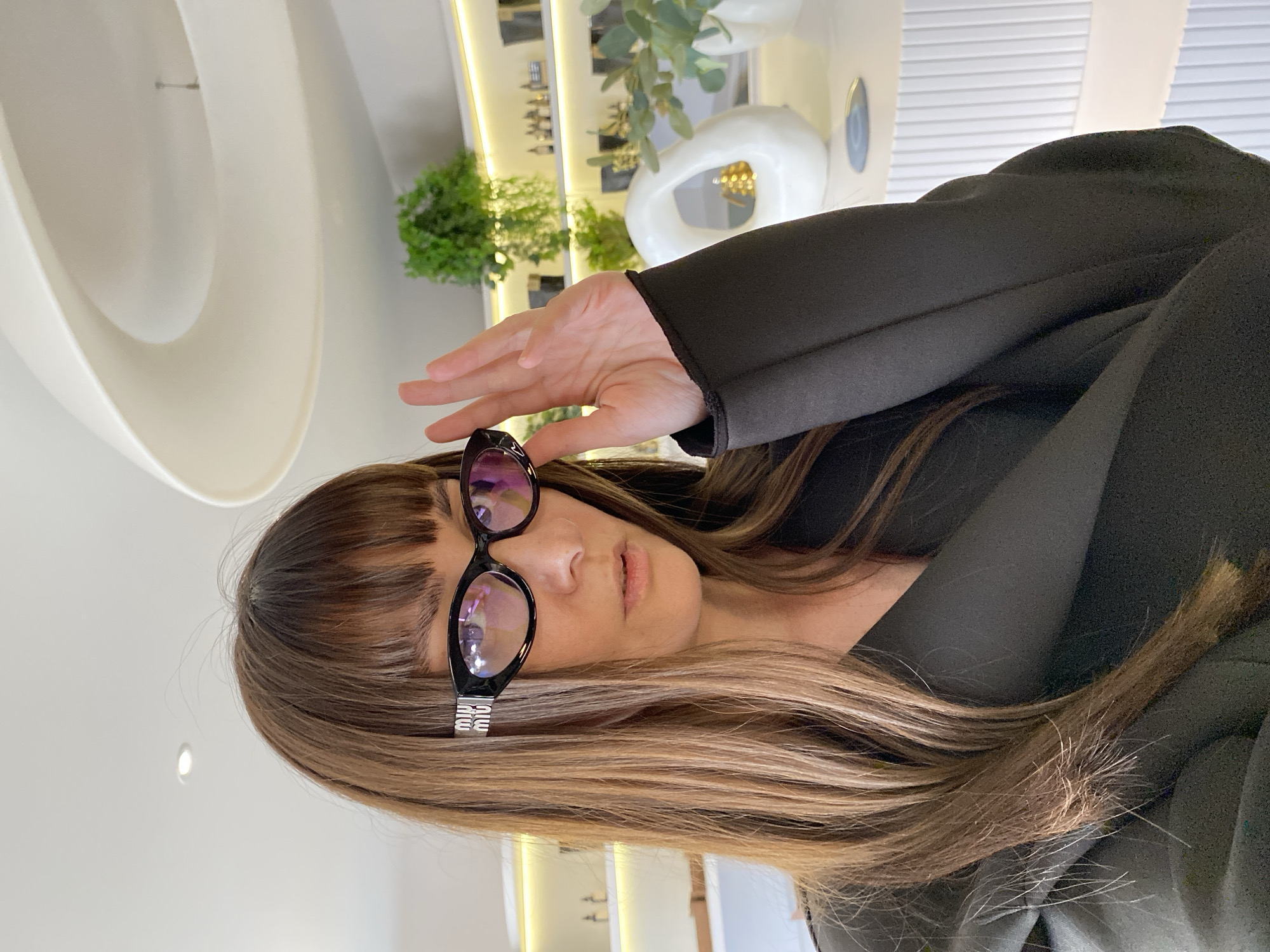 A woman with brown hair and bangs is wearing black sunglasses and a black blazer. She is holding her hand up to shield her eyes from the light. The background includes indoor plants, white decorative objects, and a shelving unit with various items. The location appears to be indoors, but it is impossible to verify if it is in Votkinsk, Russia, based on the image alone.