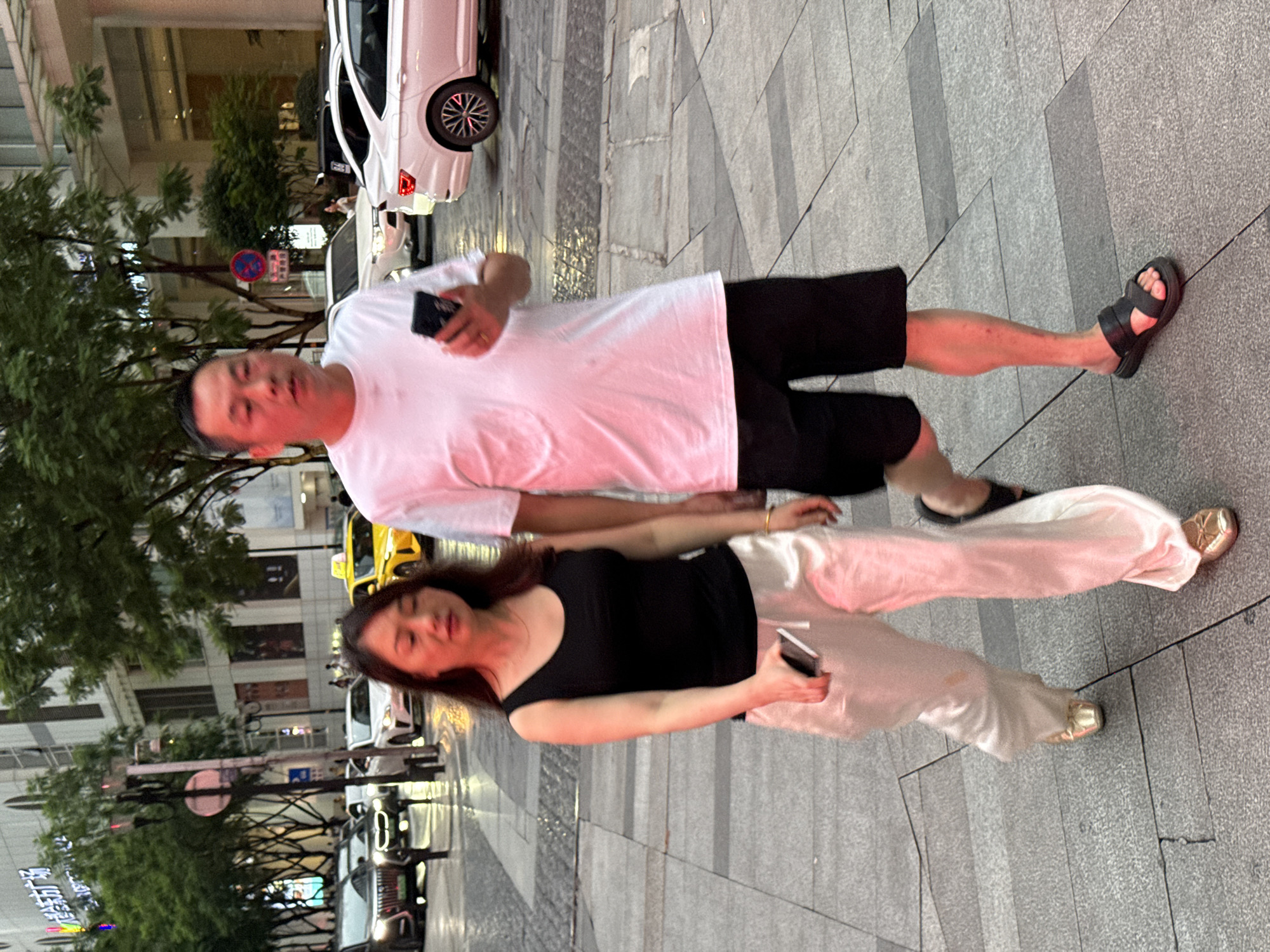 In Yuzhong District, China, a man and a woman are walking on a paved sidewalk in an urban environment. The man, on the right, is wearing a white t-shirt and black shorts, and is holding a smartphone in his right hand. The woman, on the left, is wearing a black tank top and loose, cream-colored pants, and is also holding a smartphone in her right hand. They are walking in the same direction. In the background, several cars are visible on a street, including a yellow taxi and white sedans. Buildings with trees in front of them line the street. The lighting suggests it is daytime, possibly late afternoon or early evening, with some reddish hues present. The sidewalk has a textured, grey paving pattern.