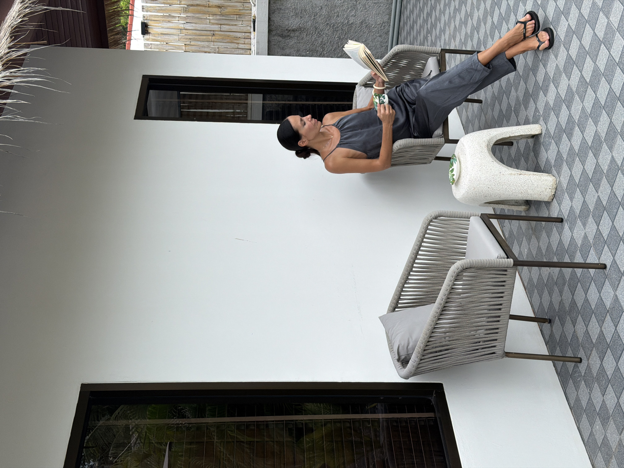 In Baan Bo Phut, Thailand, a woman is seated in a wicker chair, reading a book and holding a mug. She is wearing a dark grey sleeveless jumpsuit and black flip-flops. The chair is positioned on a grey tiled patio, next to a white wall with a dark-framed window. To her left, another wicker chair with a grey cushion is visible. A small, white, stone-like stool with a green-patterned coaster sits between the two chairs. In the background, a bamboo fence and a thatched roof suggest an outdoor setting.