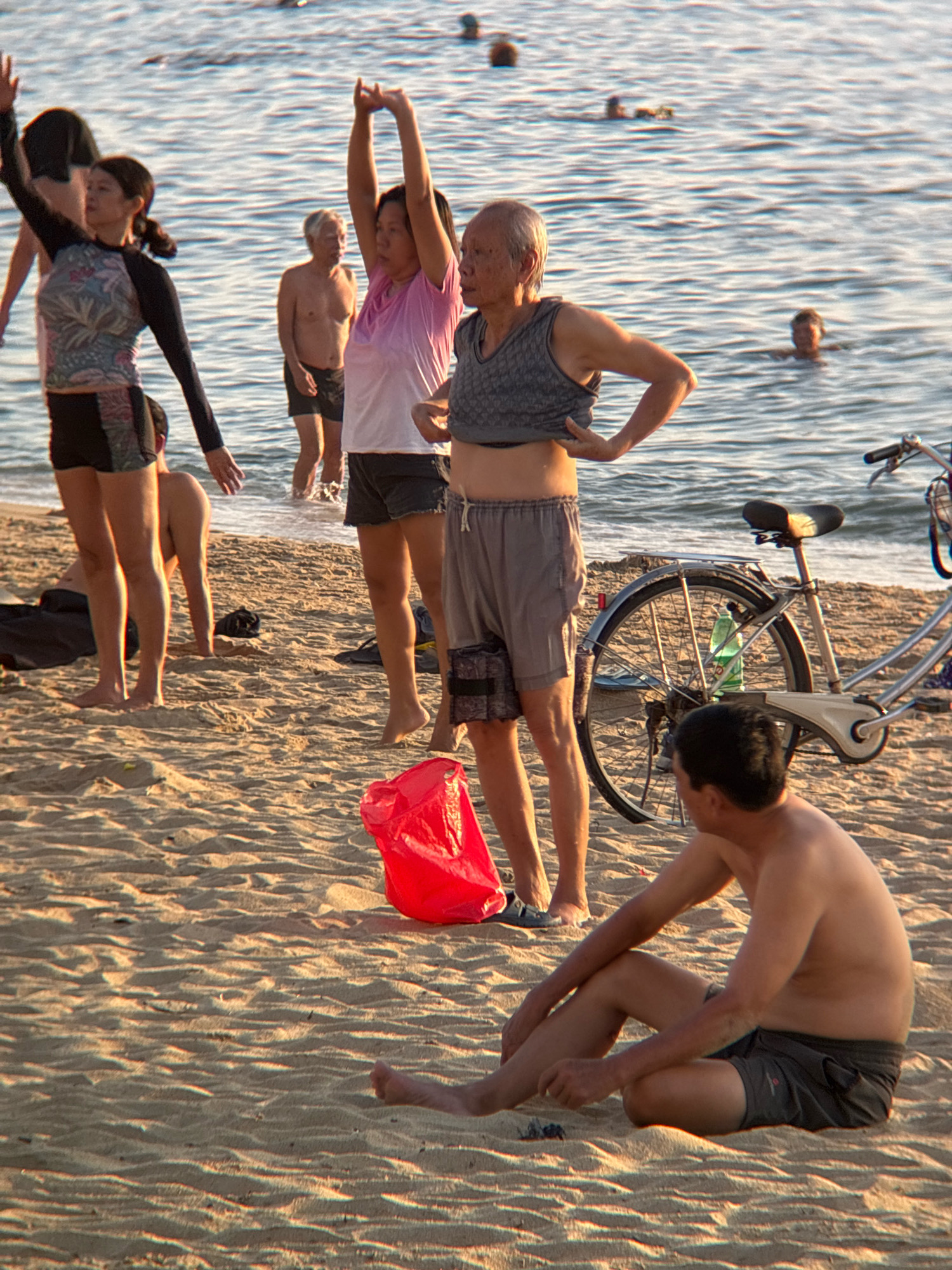 The image depicts a beach scene in Nha Trang, Vietnam, with several people engaged in various activities. In the foreground, a man sits on the sand, looking to the right. Behind him, a bicycle stands on the beach, with a green water bottle attached to the frame. To the left of the bicycle, a woman in a grey tank top and shorts is standing, adjusting her clothing. Beside her, another woman in a pink t-shirt and dark shorts is also standing, with her arms raised. Further left, a woman with dark hair and a rash guard is standing with her arms raised. In the background, several people are visible in the water, swimming or wading. The beach itself is sandy, with gentle waves lapping at the shore. The lighting suggests it is either early morning or late afternoon.