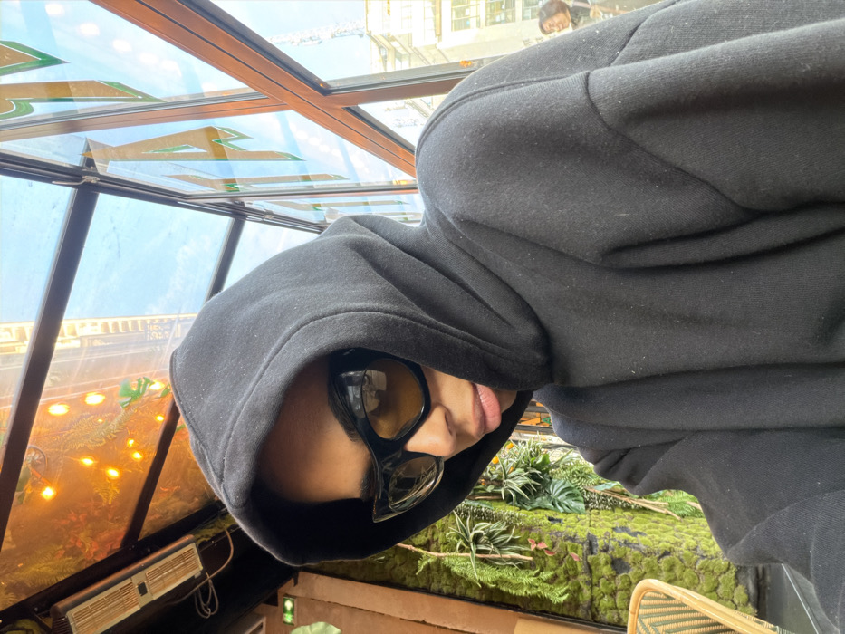 A person in a black hoodie with the hood up is wearing dark sunglasses. The person is sitting inside a structure with windows. Outside the windows, there are plants and moss. The person's head is tilted to the side. Sunlight is streaming through the windows. There is a partial view of a wicker chair.