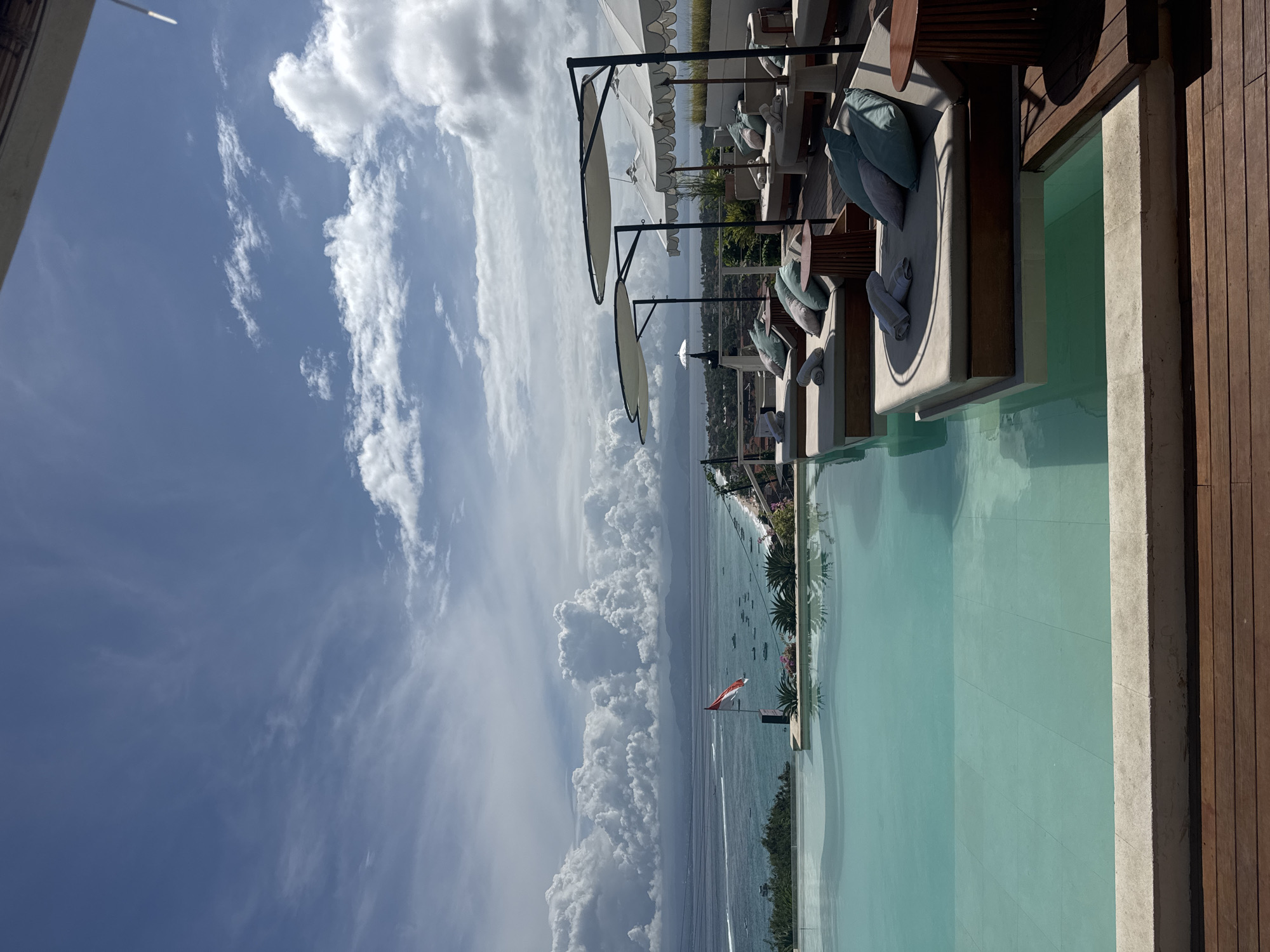 The image depicts a rooftop infinity pool overlooking the coast of Nusa Penida, Indonesia. The clear blue water of the pool merges with the ocean and sky, creating a seemingly endless vista. Several lounge chairs with white cushions and beach umbrellas are arranged along the pool's edge. The Indonesian flag is visible in the distance. The sky is filled with white, fluffy clouds. The overall scene evokes a sense of luxury and tranquility.