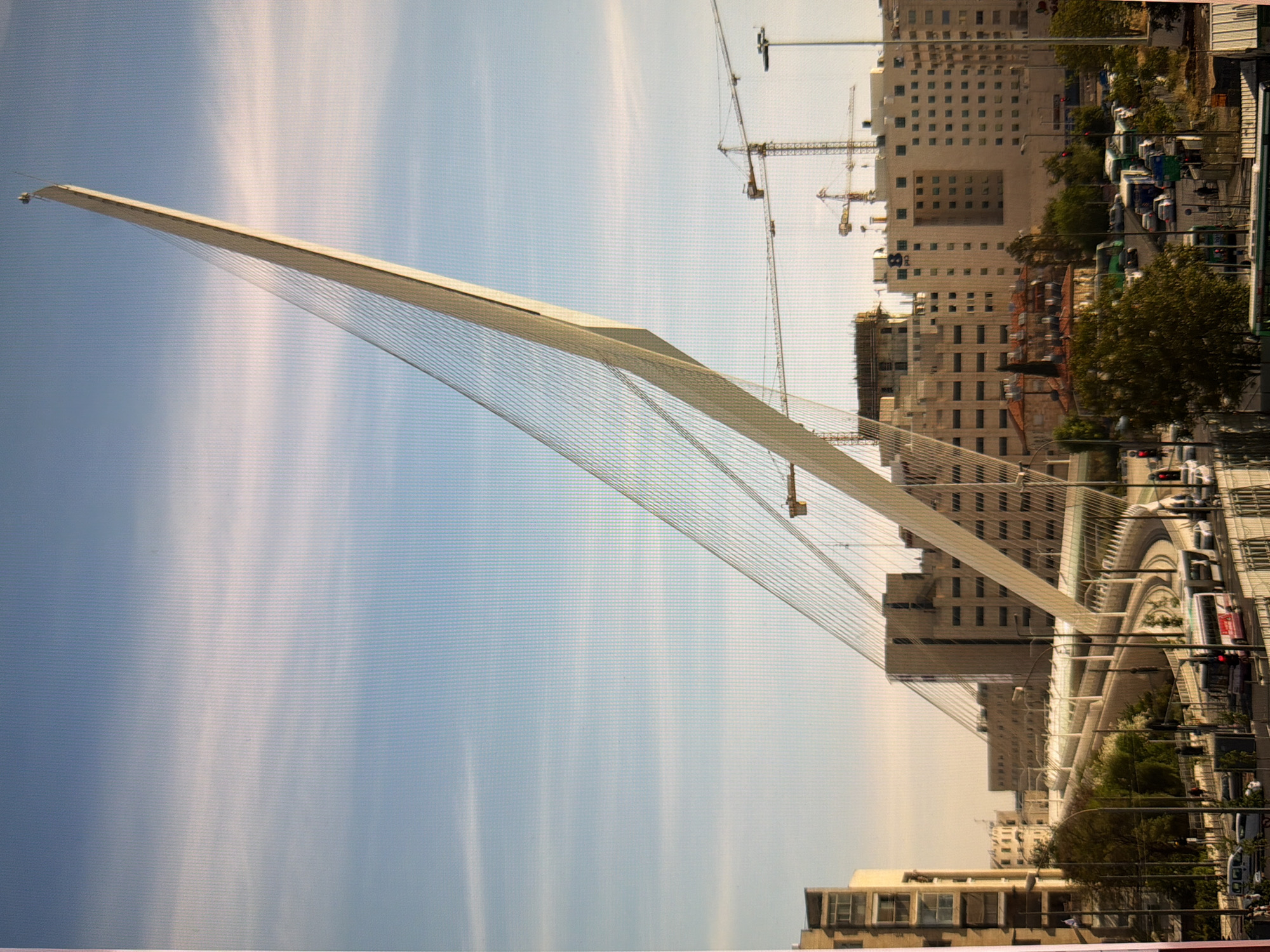 The image shows a modern cable-stayed bridge with a tall, leaning pylon, set against a blue sky with some scattered clouds. The bridge's cables are prominently visible, fanning out to support the bridge deck. In the background, there are several multi-story buildings, some appearing to be under construction with cranes visible. Below the bridge, there is a road with traffic, including buses and cars. Trees and streetlights are also present. The scene depicts an urban environment with a focus on the architectural structure of the bridge. The location is not Kyiv, Ukraine.