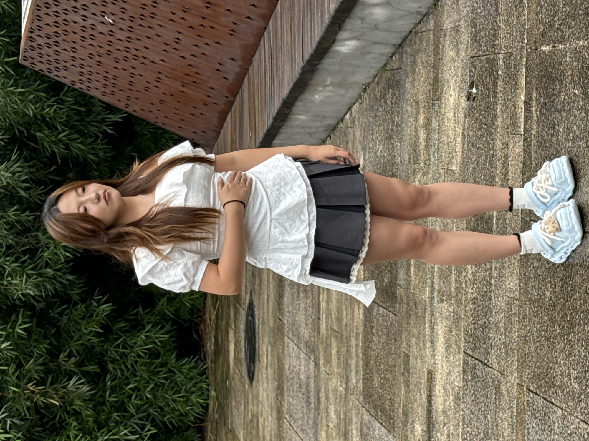 A young East Asian woman stands outdoors on a wet, paved pathway. She has long, wavy brown hair and is wearing a white, off-the-shoulder top with ruffled sleeves and a layered ruffled hem, over a dark gray pleated skirt with a lace trim. She has one hand on her stomach and is looking to her right. She is wearing white socks with black lace trim and light blue clogs with white laces. In the background to the left, there are dense green bamboo stalks. To the right, there is a wooden bench with a patterned, rust-colored metal structure behind it. The paving stones are a mix of light and dark gray. The scene appears to be in a park or public outdoor space within Yuzhong District, China.
