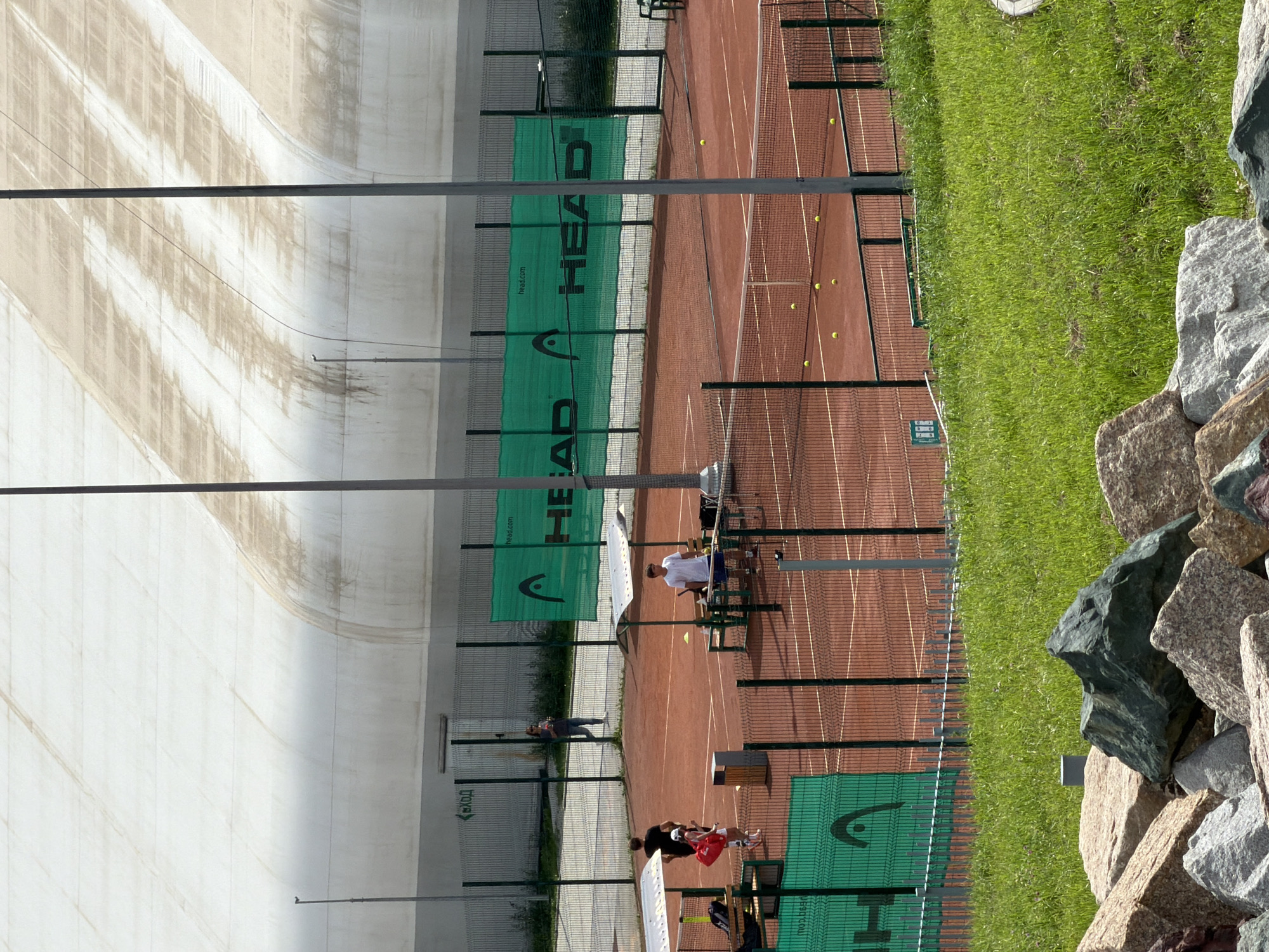 The image depicts an outdoor tennis court complex with a large, white, domed structure in the background. The court is made of red clay and has tennis nets. Several people are present on and around the courts. On one court, two individuals, one appearing to be an instructor, are interacting with a person holding a tennis racket. Another person is standing further away, holding a drink. On a different court, a young male in a white t-shirt and blue shorts is holding a tennis racket and standing near a rack with tennis balls and equipment. Tennis balls are scattered across the courts. Green banners with the "HEAD" logo are visible along the fences surrounding the courts. In the foreground, there is a grassy slope with large rocks. The environment suggests a recreational or training facility. The signage in Russian ("ВХОД" which means "ENTRANCE") indicates a location in Russia. The specific location in Yekaterinburg, Russia, is the "Tennis Club Yekaterinburg," also known as the "Tennis Club of the Yekaterinburg Forest-Park Association," situated at the address ulitsa Vostochnaya, 4G.