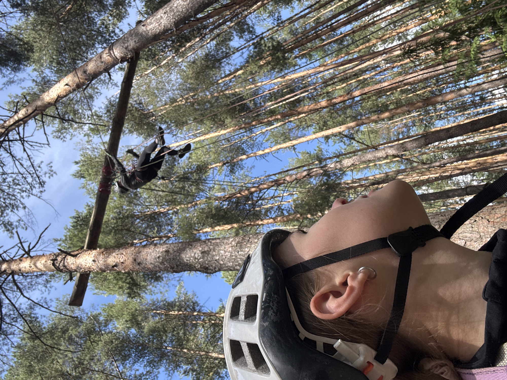 In a forest setting, likely within the city of Chaikovsky, Russia, a person wearing a white helmet and dark clothing is being suspended mid-air from a horizontal wooden beam supported by a large tree. The person appears to be engaged in an activity like zip-lining or abseiling, with ropes visible connecting them to the beam. The foreground is dominated by a close-up of another person's face and helmet, looking upwards towards the suspended individual. The background consists of a dense forest of tall, slender pine trees under a clear blue sky.