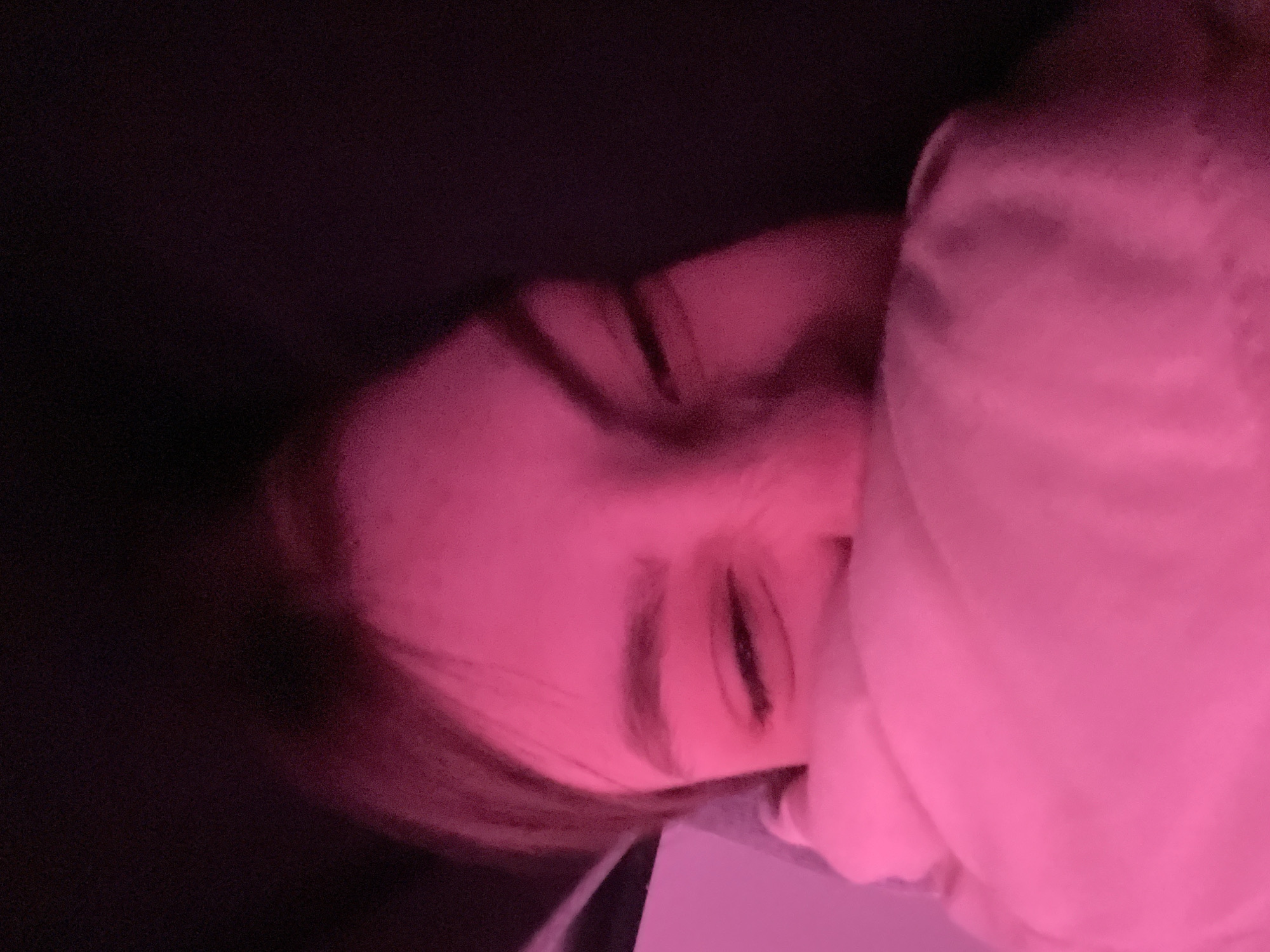 The image shows a person's face partially obscured by a pink-lit object. The person is lying down, and the face appears to be in a relaxed or sleeping state, with closed eyes and relaxed facial muscles. The lighting is predominantly pink, which may distort the true skin tone and colors of the objects. The background is dark. The location cannot be determined from this image.
