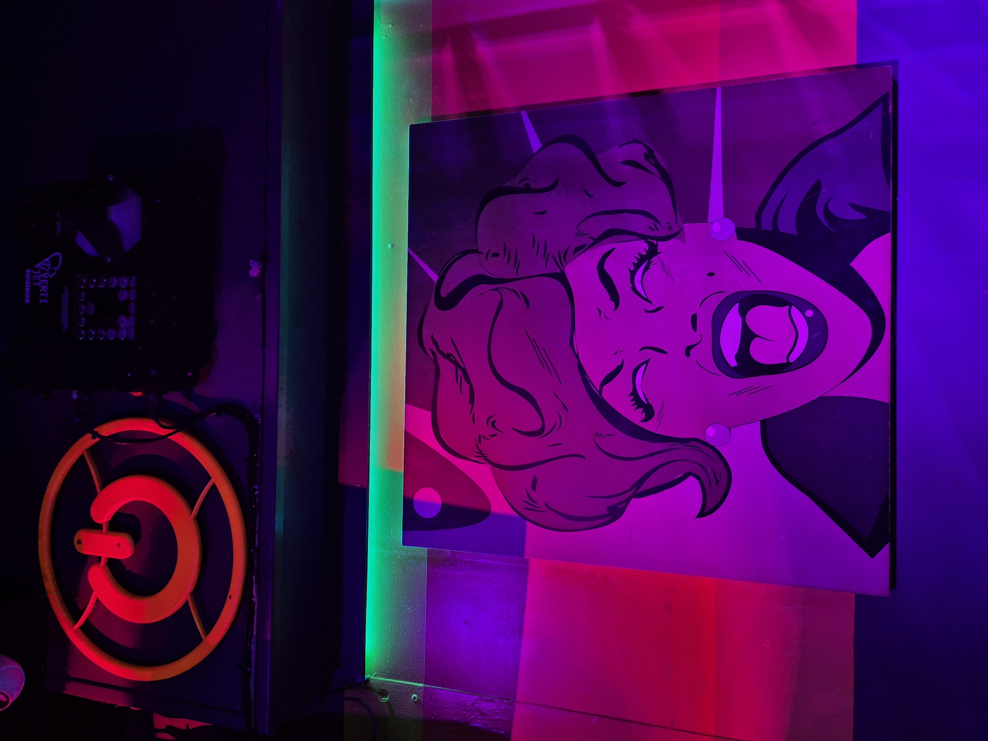 The image shows an interior space with a purple and pink neon color scheme. A piece of art depicting a woman with fangs is mounted on the wall, illuminated by the pink and purple lighting. To the left of the artwork is a black device with a circular orange fixture attached, possibly a ventilation or cooling system. A green light is seen at the edge, indicating a decorative neon strip. The location is Cardiff, United Kingdom.