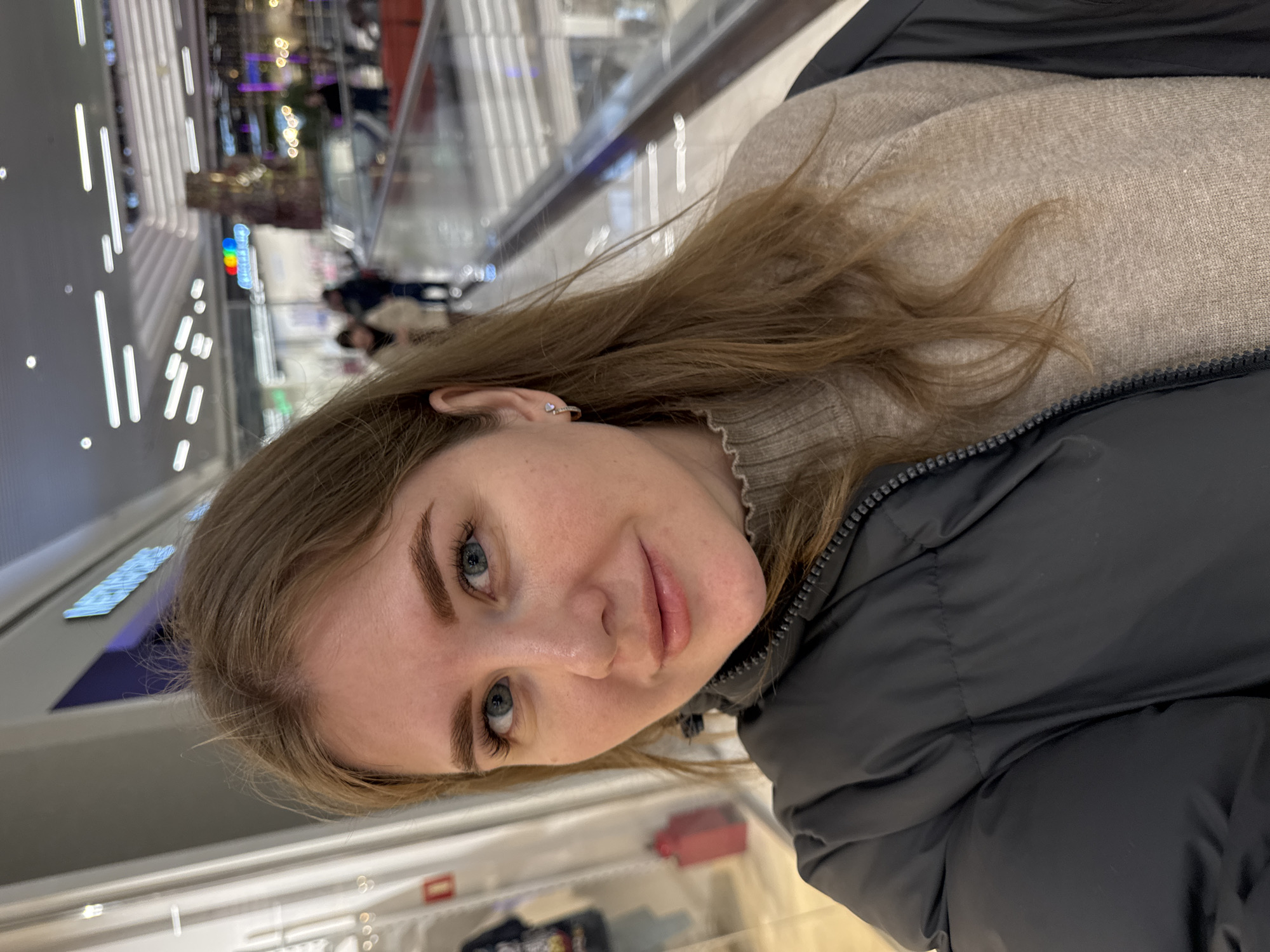 The image is a selfie of a woman inside a shopping mall. The woman has fair skin, light brown hair, and blue eyes. She is wearing a beige sweater and a black puffer jacket. The background shows a modern shopping mall interior with multiple levels, glass railings, and illuminated ceiling lights. In the blurred distance, there are store signs and other shoppers. The location is likely a shopping mall in the city of Perm, Russia, although the exact identity of the mall is not discernible from the image alone.