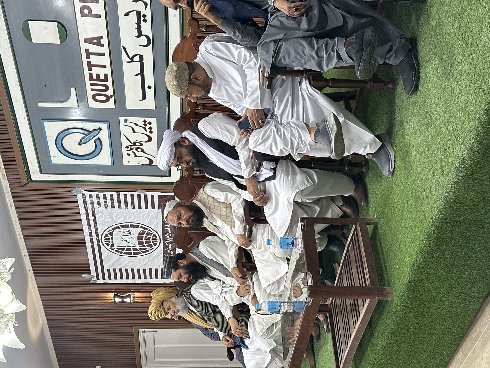 A group of six adult men are seated in a row of chairs on what appears to be artificial turf. The men are wearing traditional Pakistani clothing, with some wearing turbans or skullcaps. A table runs along the front of the chairs, with several bottles of water on it. Behind the men is a wooden wall. A banner hangs on the wall with a logo in the center and script above and below. Above the banner is a sign that reads "QUETTA PRESS CLUB" in English and Urdu. The sign also includes a logo and some additional script in Urdu. The location is likely the Quetta Press Club in Quetta City Tehsil, Pakistan.
