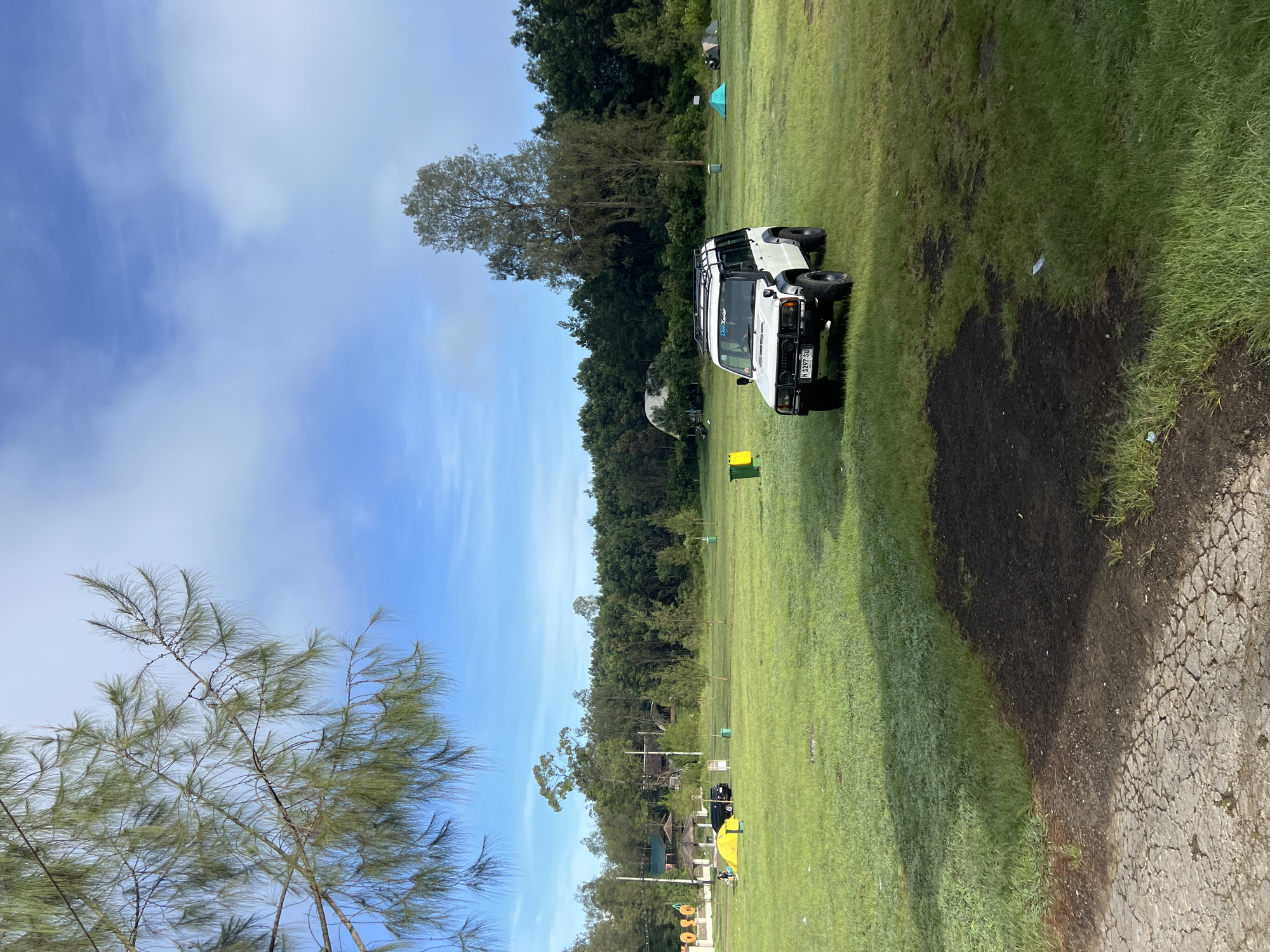 A white SUV with a roof rack is parked on a grassy field in an outdoor location near Unknown, Indonesia. The sky is blue with scattered clouds. A tree with sparse foliage stands to the left of the frame. To the right of the SUV is a darker patch of grass. In the background, there's a dense tree line. A portion of cracked pavement is visible in the lower right corner. A yellow trash can sits near the SUV. Tents and structures are seen in the distance amidst the trees.