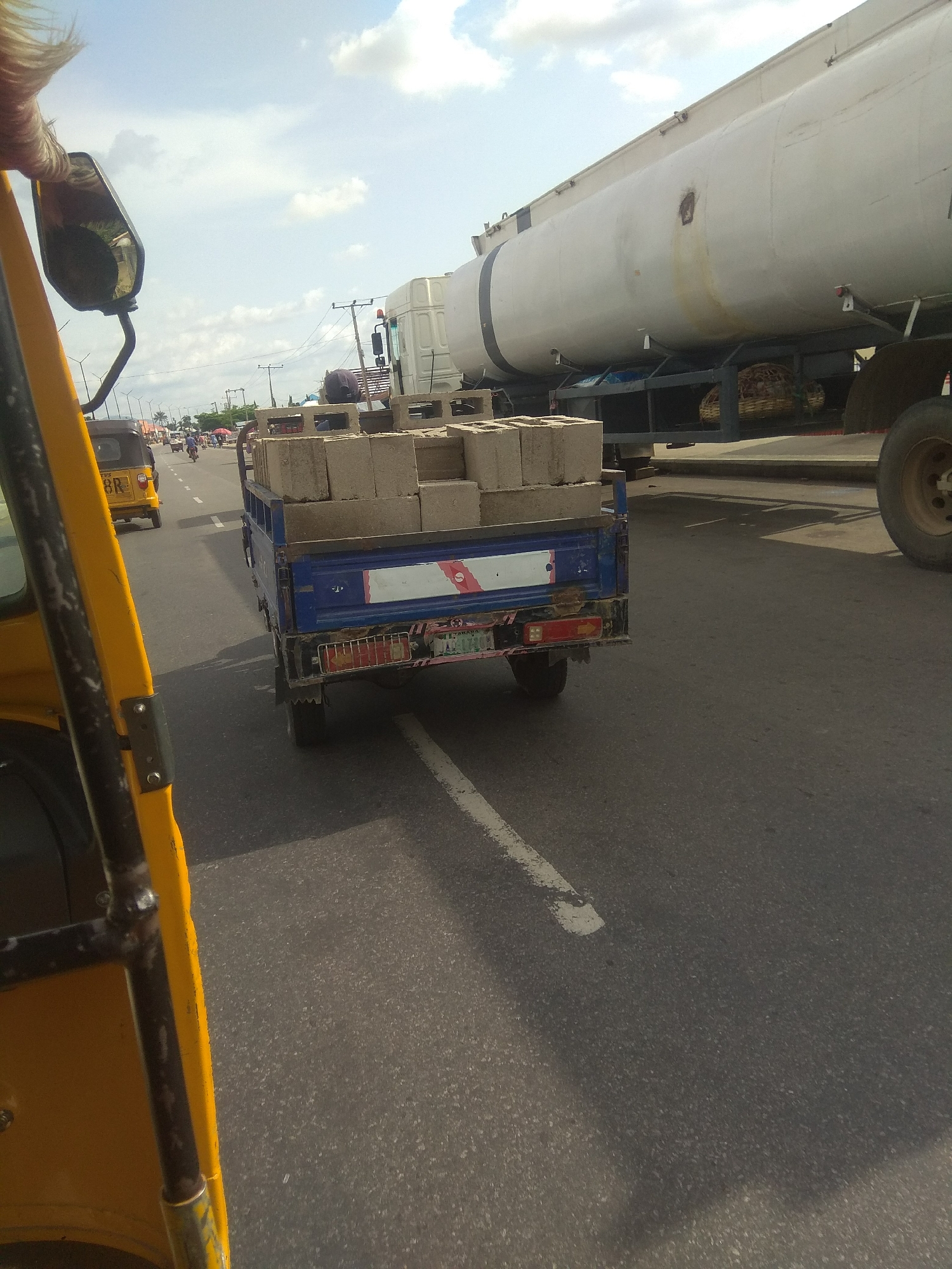 In Jalingo, Nigeria, a bright yellow auto-rickshaw navigates a road, its perspective dominating the foreground. The vehicle's interior and side are visible, hinting at the driver's viewpoint. Further down the road, a small blue truck is laden with concrete blocks, its cargo secured for transport. Behind the truck, a large white tanker truck is parked or moving slowly, its cylindrical body stretching across the frame.
The scene appears to be set during daylight hours, under a partly cloudy sky, suggesting a warm day. The road is paved and marked with a white line, indicating a traffic lane. Other vehicles, including what appears to be another auto-rickshaw and a few motorcycles, are visible in the distance, suggesting moderate traffic. The presence of utility poles and buildings on the side of the road points to an urban or semi-urban environment.
The overall atmosphere seems ordinary, depicting a typical scene of transportation and commerce on a Nigerian road. There are no overtly noticeable emotional cues from any visible subjects. The visible text on the yellow auto-rickshaw includes "BR" on the rear license plate. The blue truck also has some markings, including a red and white stripe and what looks like a license plate at the bottom rear. The tanker truck has some faded markings and a dark stain on its side.
