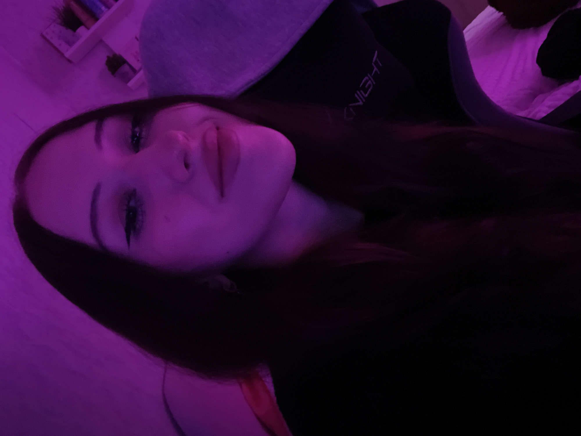 An individual, presumed female, is depicted in a close-up indoor portrait in the city of Votkinsk, Russia. The subject is angled slightly to the right, looking directly towards the camera, illuminated primarily by a strong magenta/purple light source from the front-left. The individual has long, dark hair with a center part, visible winged eyeliner, and lipstick on slightly parted lips, indicating a subtle smile. They are wearing a dark-colored garment. Behind the subject, a dark gaming chair is visible, with the word "KNIGHT" partially visible in white text on the chair's backrest, oriented to the right of the subject. A light-colored, possibly grey, padded surface is visible to the upper left of the subject. In the upper right background, a white shelving unit contains several small objects, including what appears to be a small potted plant and other decorative items.