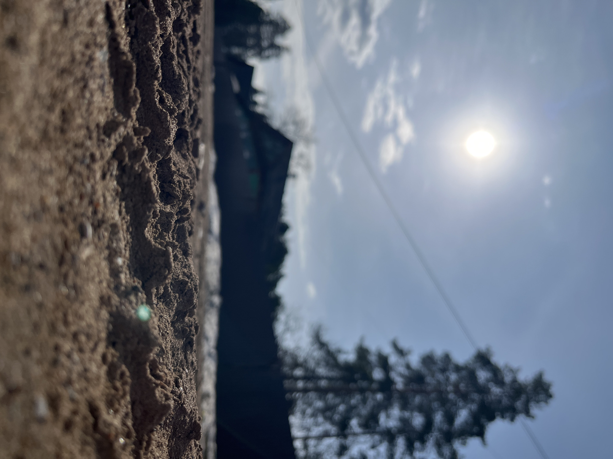 The image, taken in Chaikovsky, Russia, presents a blurred, tilted perspective. The foreground features a close-up of a rough, earthy-brown surface, likely a wall or embankment, showing detailed texture. The background, heavily out of focus, includes a bright sun in a blue sky with scattered clouds, blurred silhouettes of trees, and a dark, undefined structure resembling a building. An overhead power line is faintly visible. The overall scene suggests an outdoor environment on a sunny day, observed from a low angle near a textured surface.